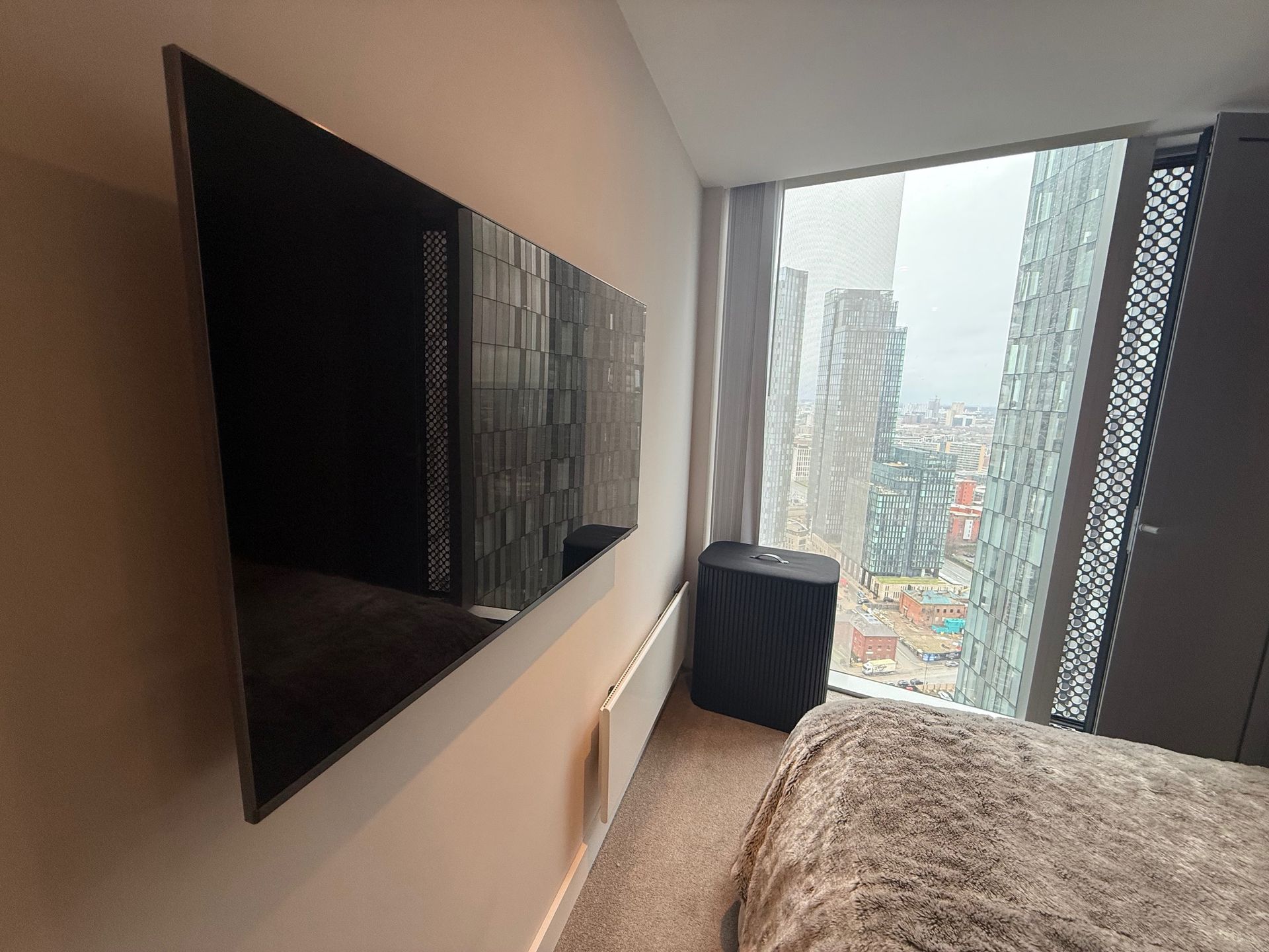 A flat-screen TV mounted on a wall. A city view is seen through the window. A bed and a cabinet are also present.