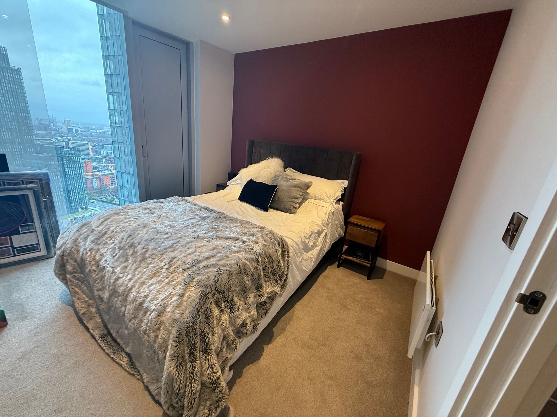 Bedroom with bed, burgundy wall, and city view through large window.