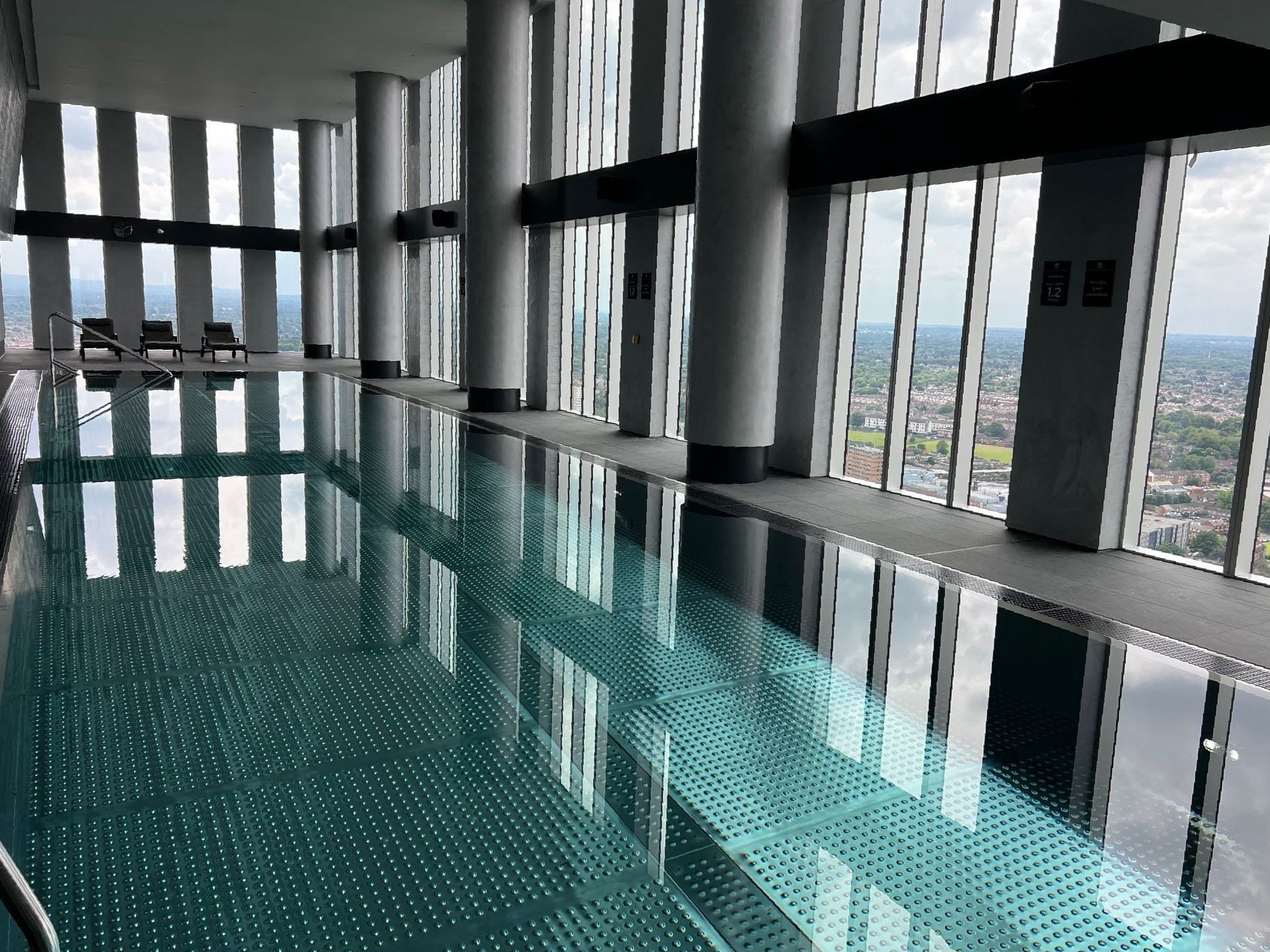 Indoor swimming pool in a high-rise building with city views. Glass windows, columns, and reflections in the water.