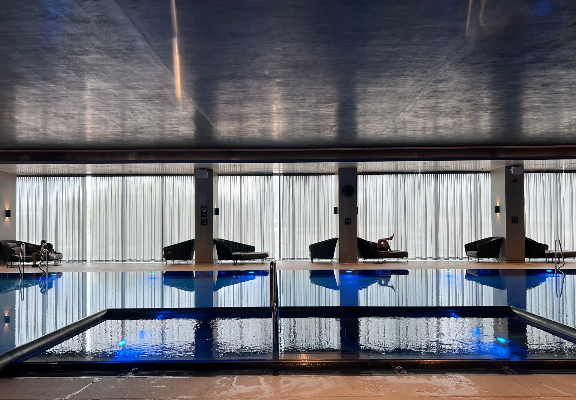 Indoor pool with blue water and lounge chairs against a white curtained wall.