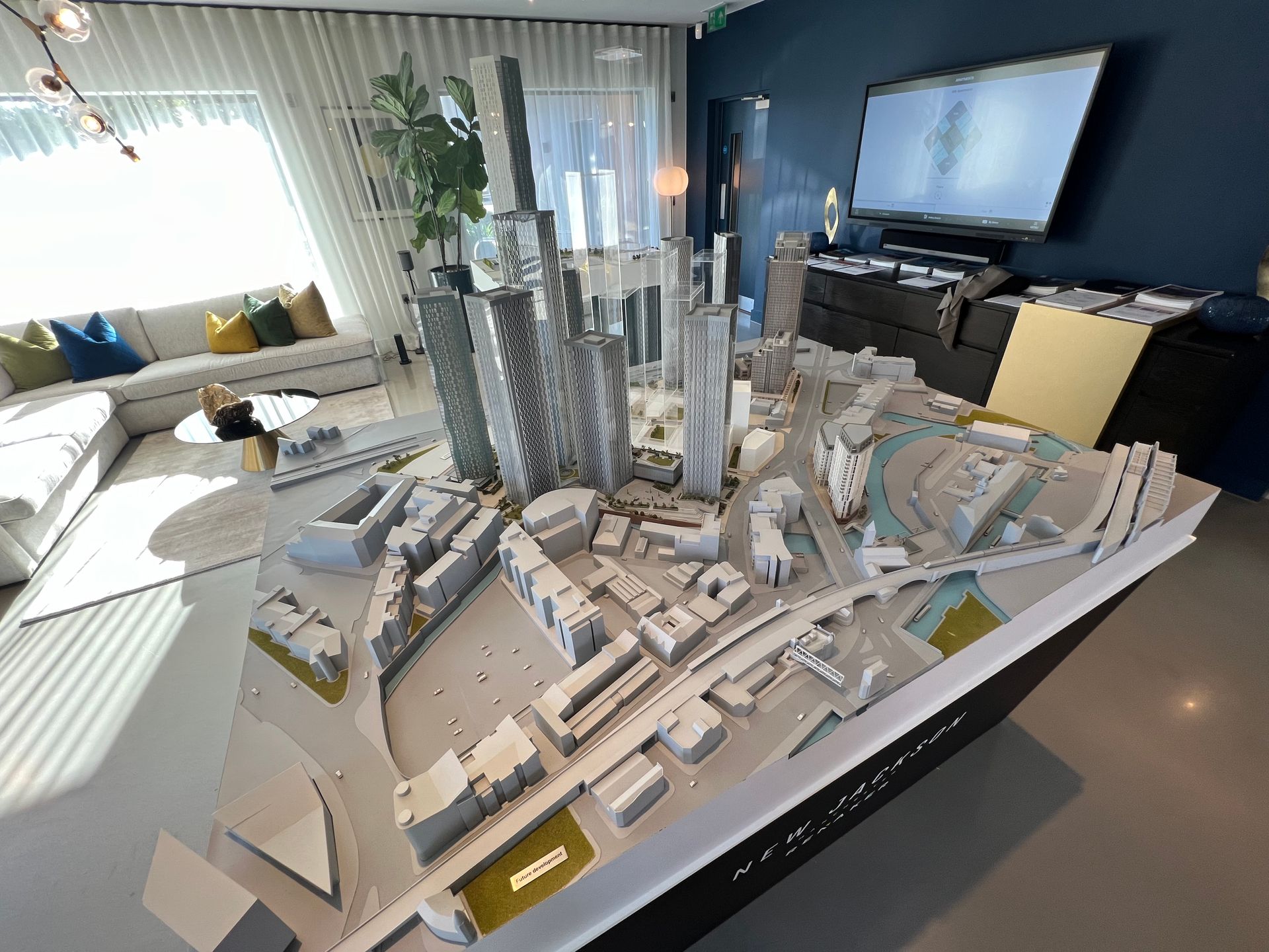 Architectural model of a city, on a table. Displayed in a room with a TV, couch, and large windows.