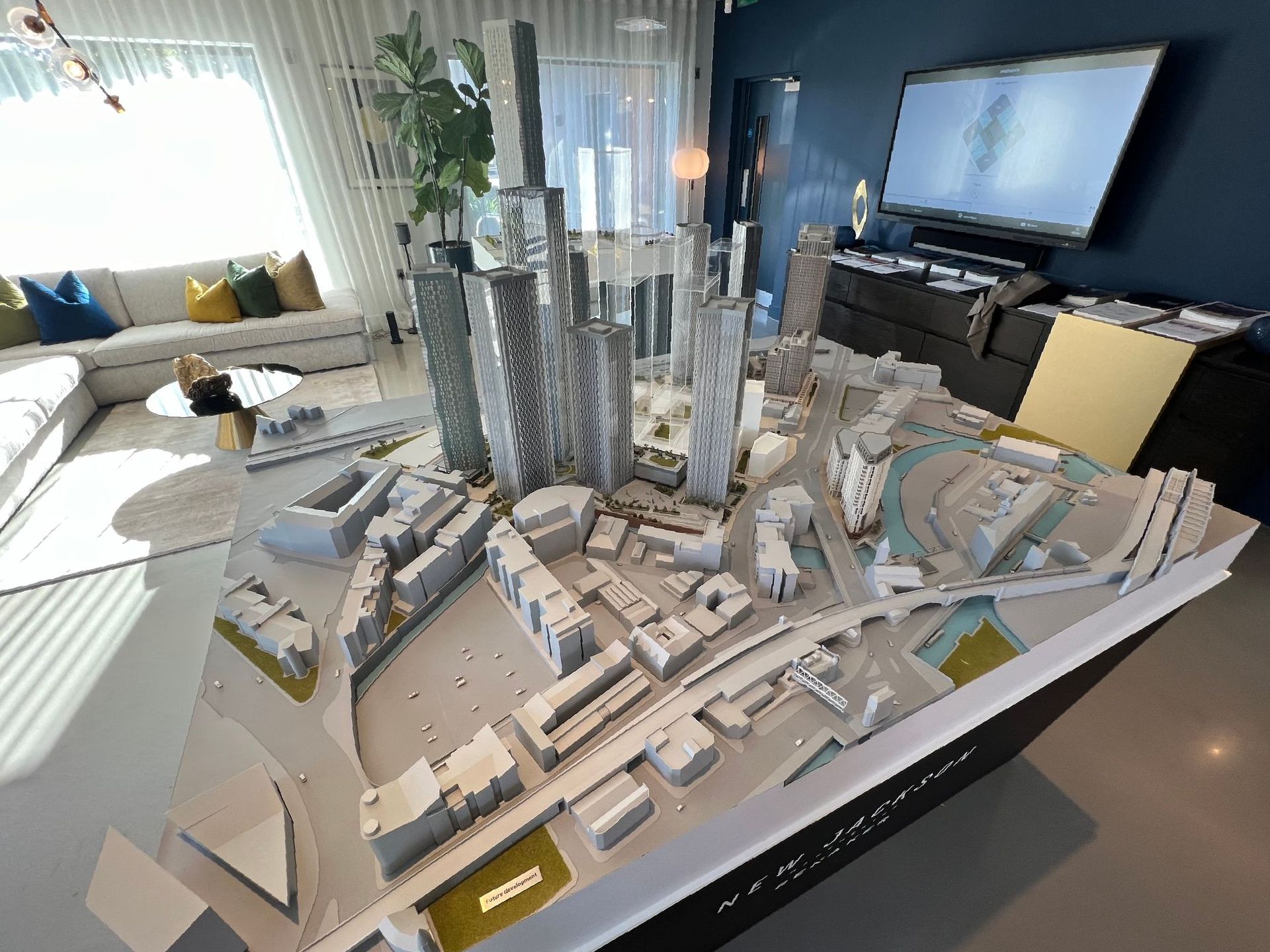 Model of a city with tall buildings, on a white table in a room with a window, couch, and TV.