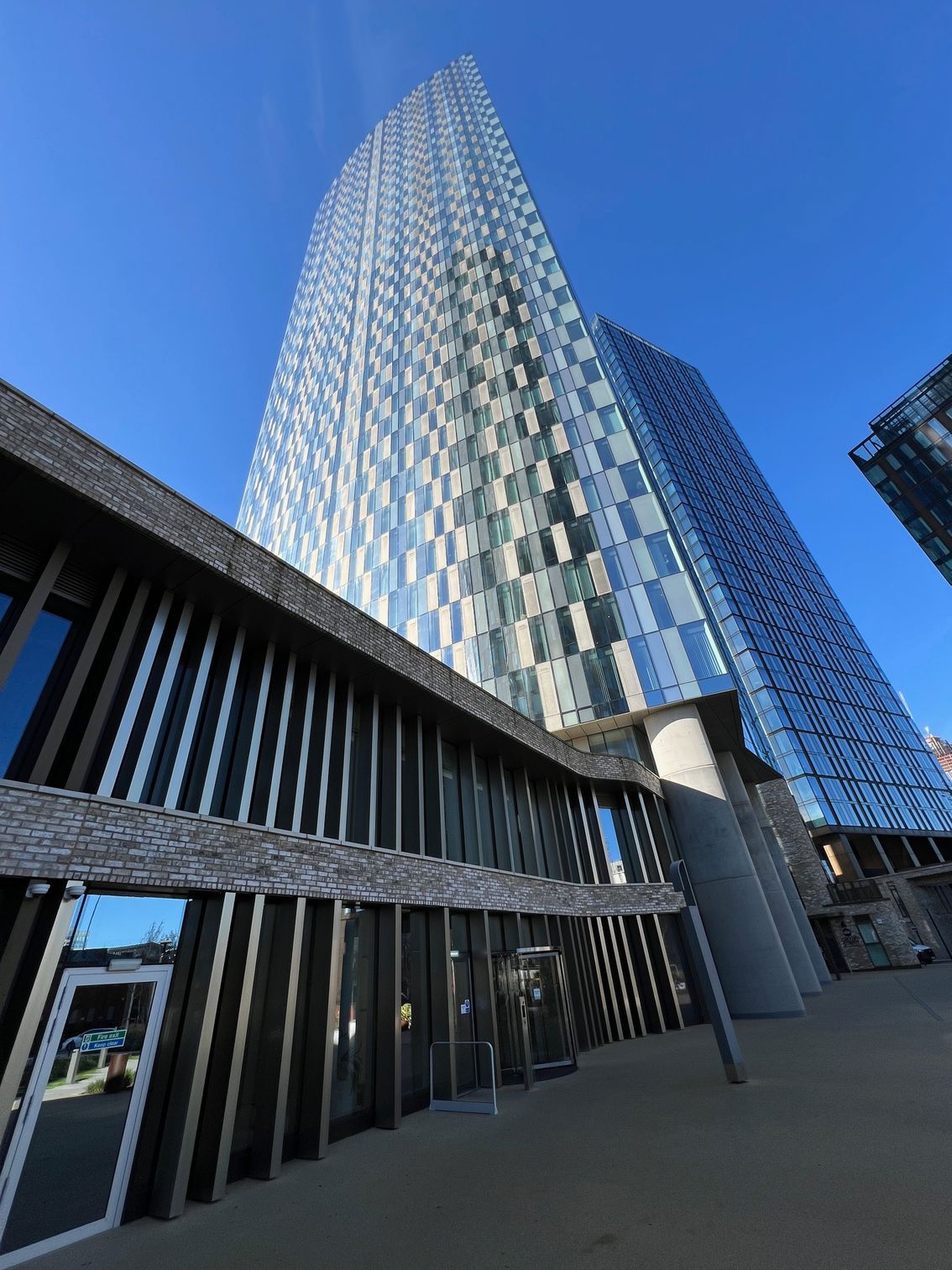 A tall, modern skyscraper with a checkered glass facade stands behind a low, wooden-paneled building; blue sky.