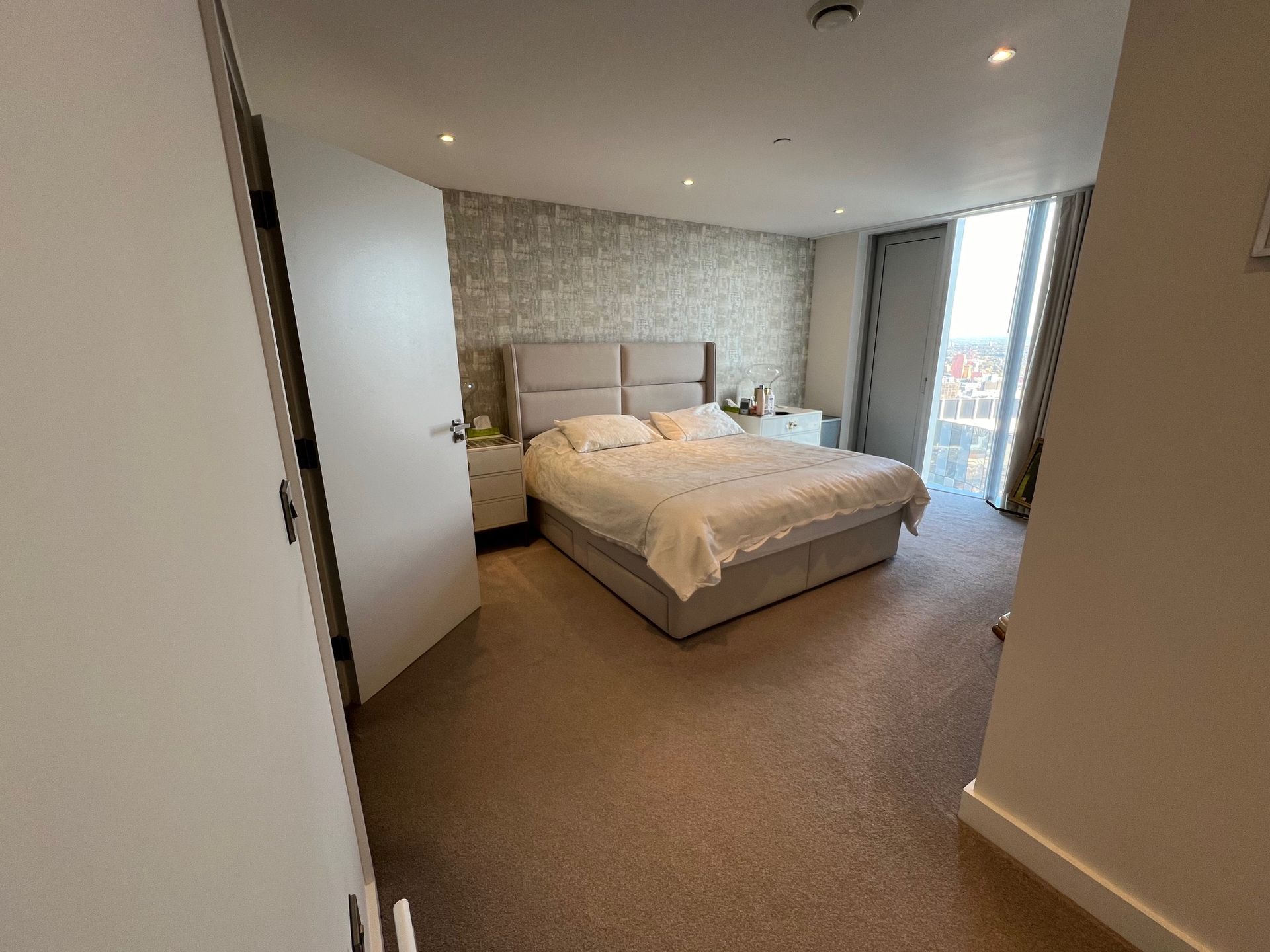 Bedroom with bed, nightstands, and window with curtains. Neutral color palette, carpeted floor.