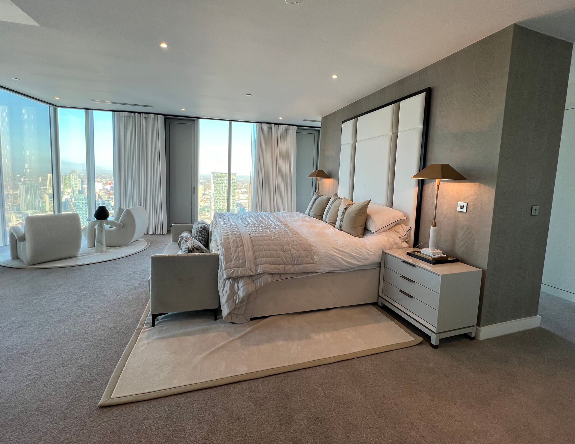 Spacious bedroom with a large bed, seating area, and floor-to-ceiling windows offering a city view.