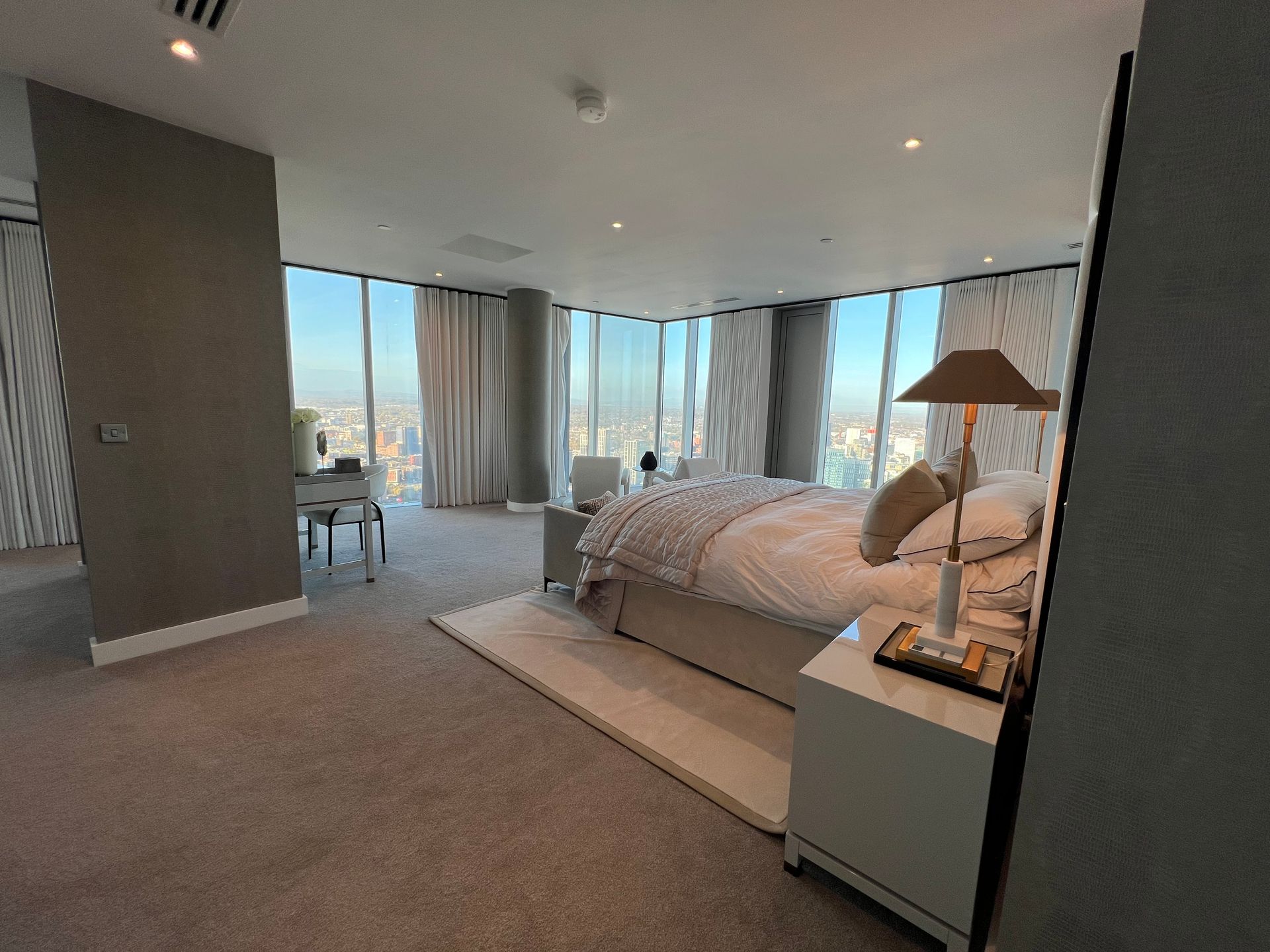 Modern bedroom with large windows, city view, bed, desk, and neutral color scheme.
