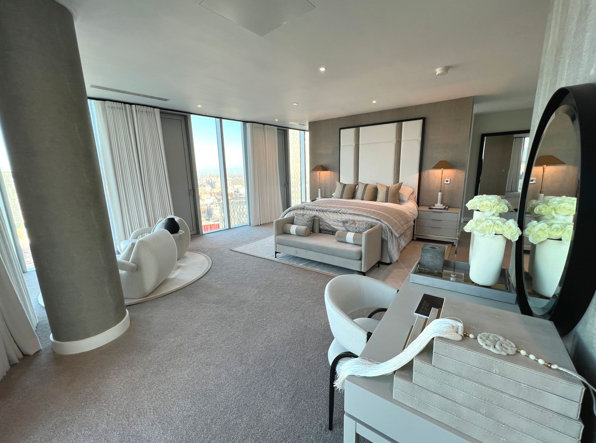 Spacious bedroom with city views, gray and white decor, large bed, vanity, and a round mirror.