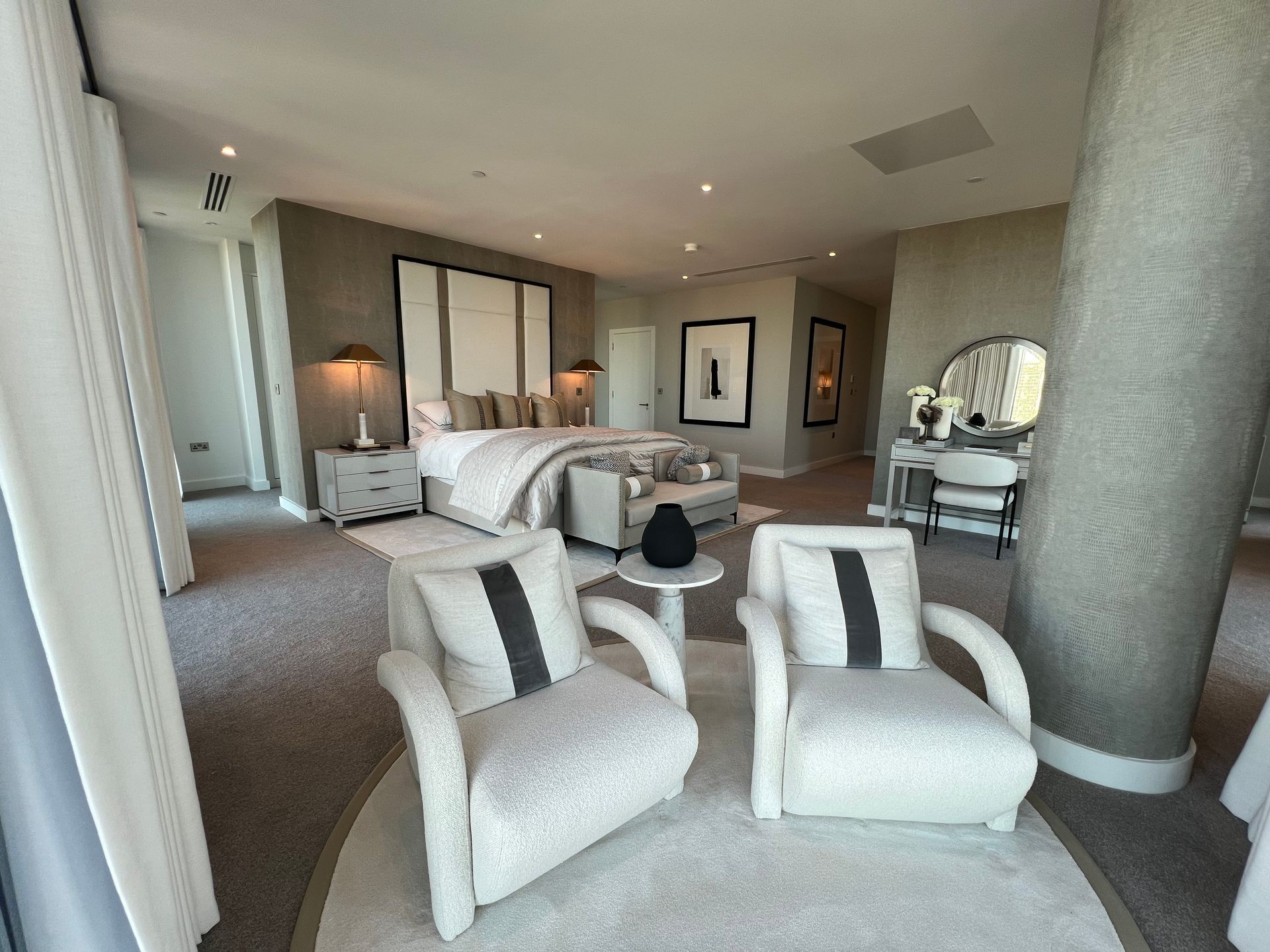 Spacious bedroom with two armchairs, bed, and vanity. Neutral tones, carpet, and a large window.