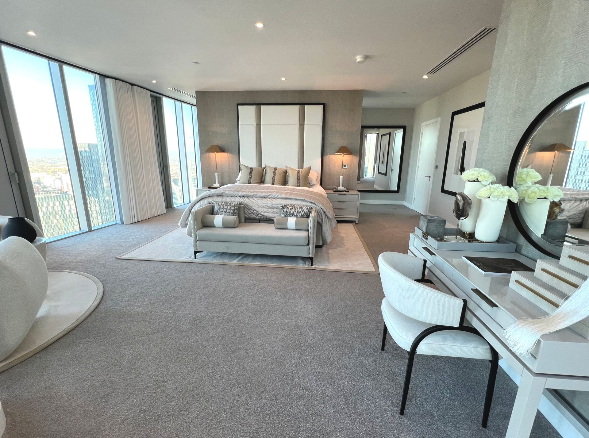 Luxurious bedroom with bed, vanity, and large windows offering city views. Neutral tones, modern decor.