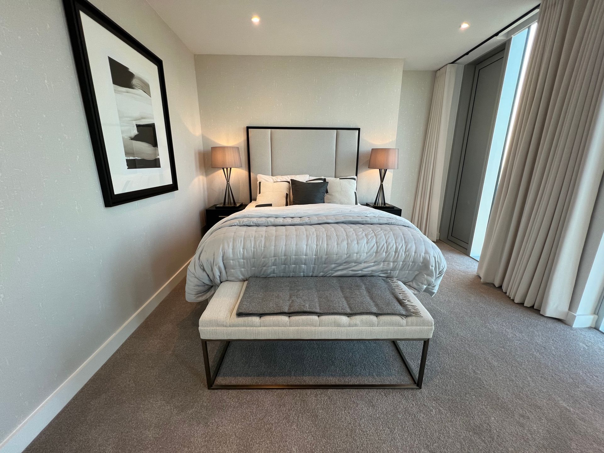 Bedroom with a bed, bench, artwork, two lamps, and a window with sheer curtains.
