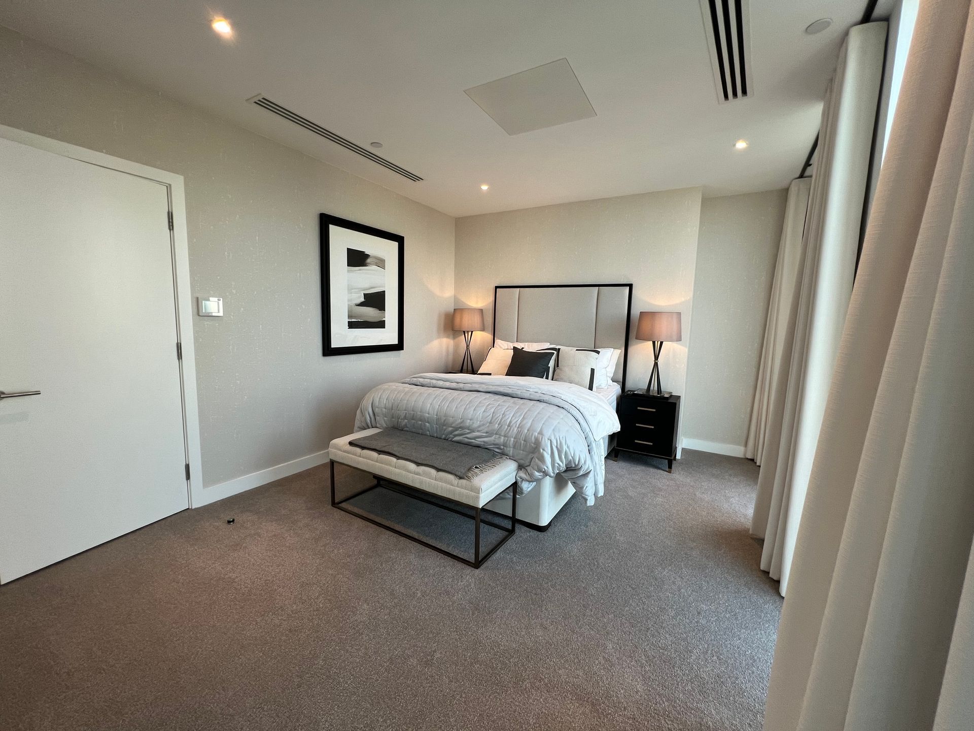 Bedroom with bed, bench, nightstands, artwork, door, and windows. Neutral colors. Carpeted floor.
