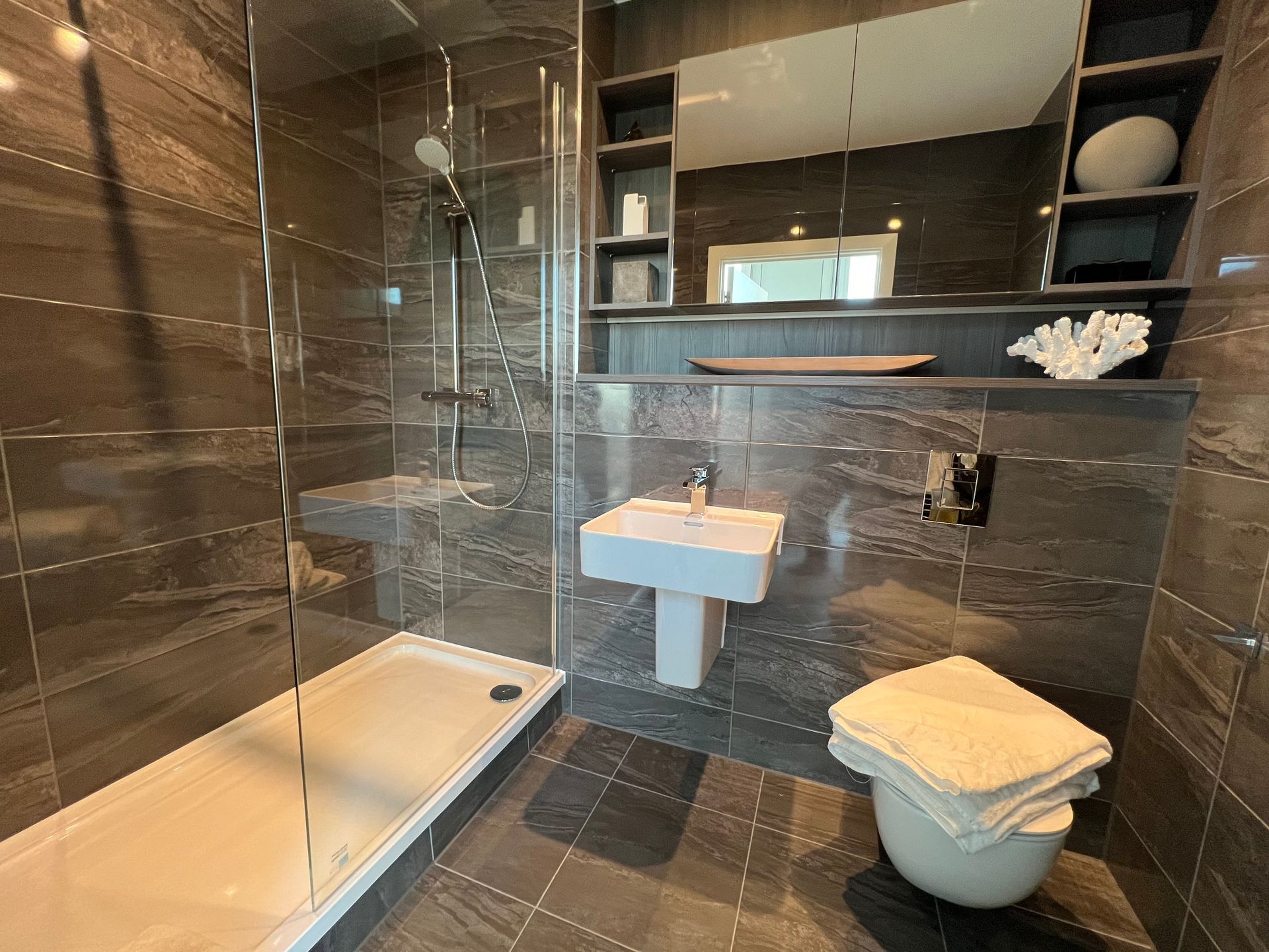 Modern bathroom with gray tiled walls, glass shower, pedestal sink, and a toilet with towels.