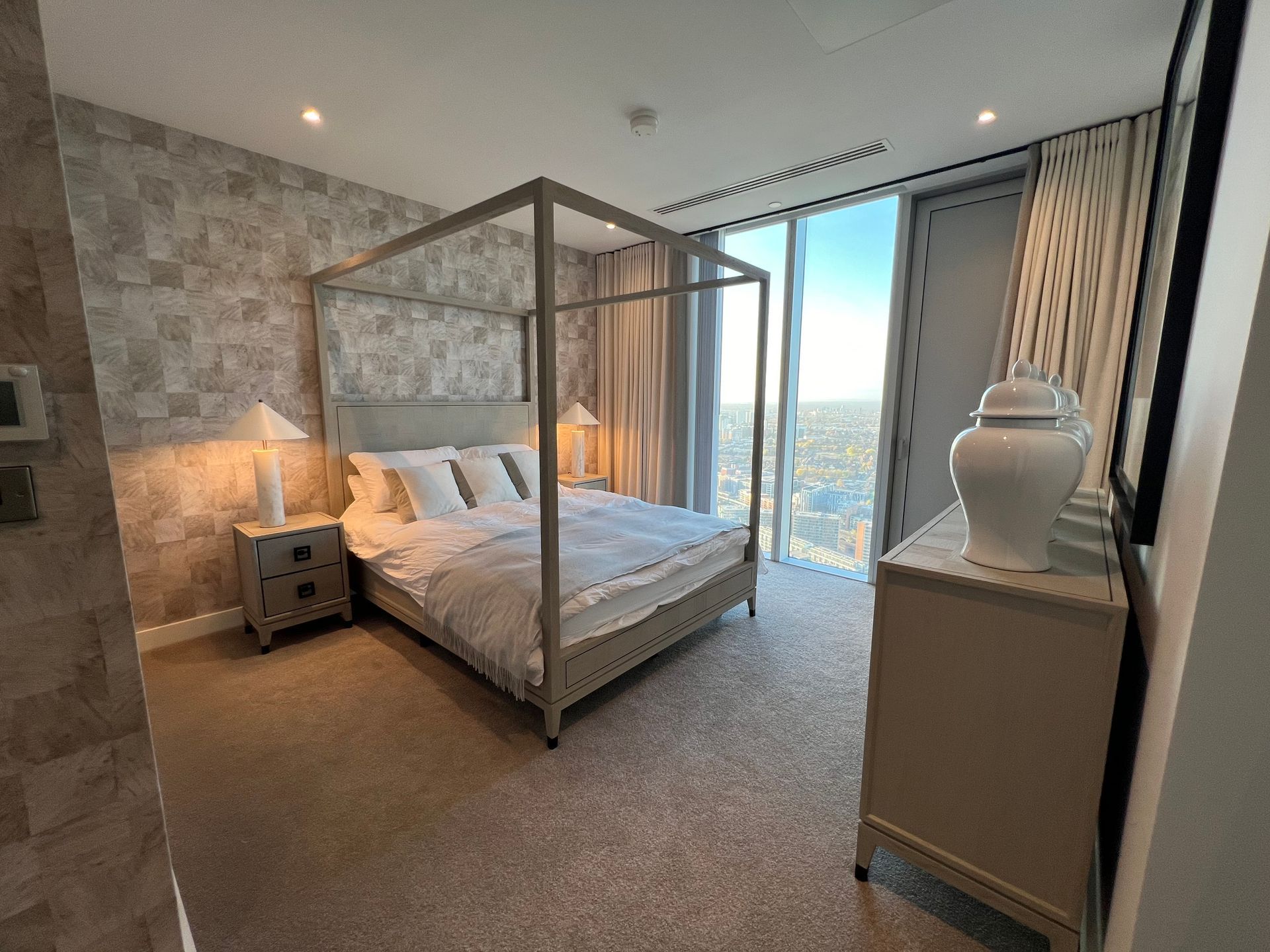 Bedroom with four-poster bed, nightstands, dresser, and large window with city view. Neutral tones, well-lit.