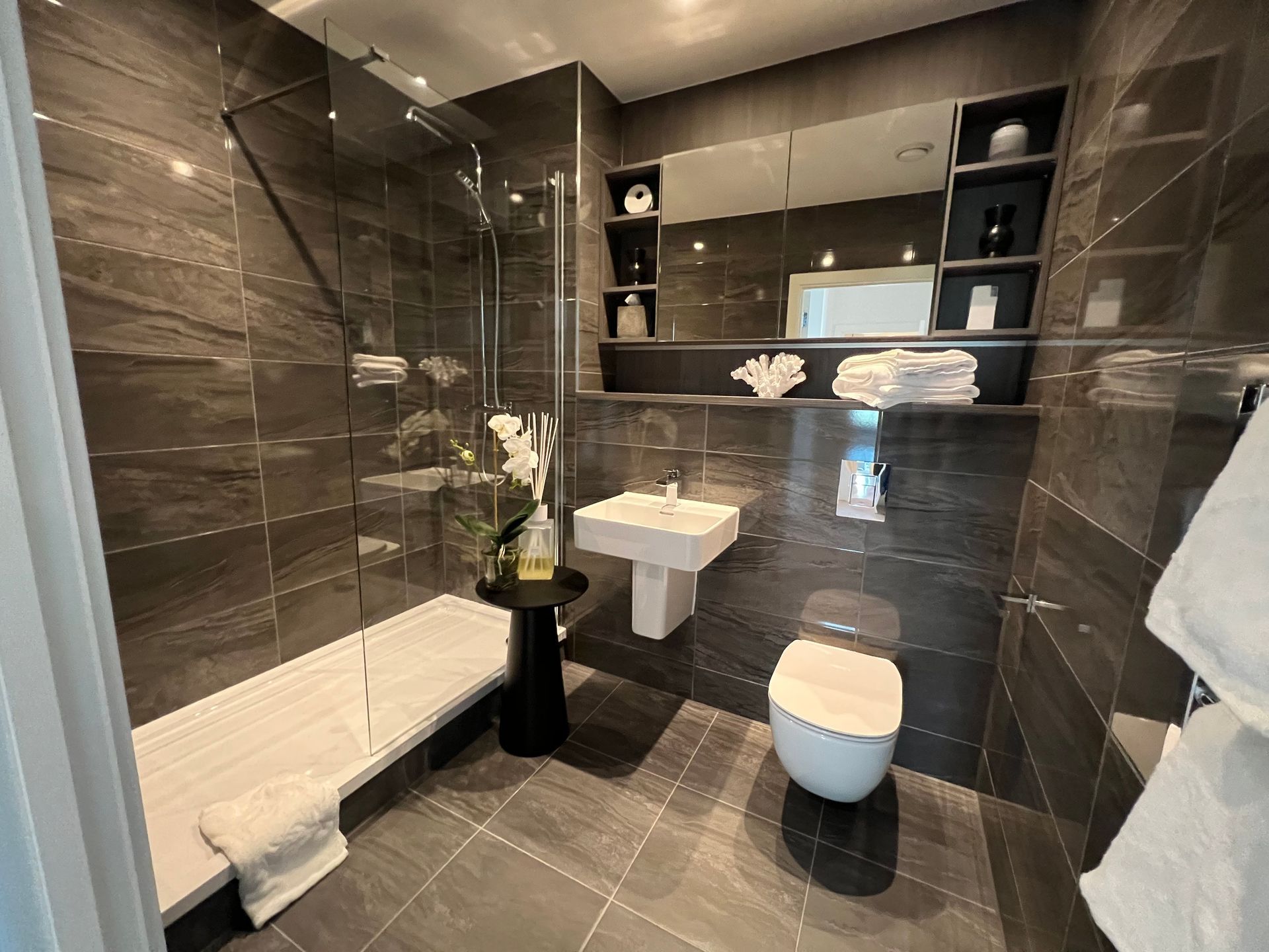 Modern bathroom with dark gray tile, glass shower, white fixtures, and a black side table.