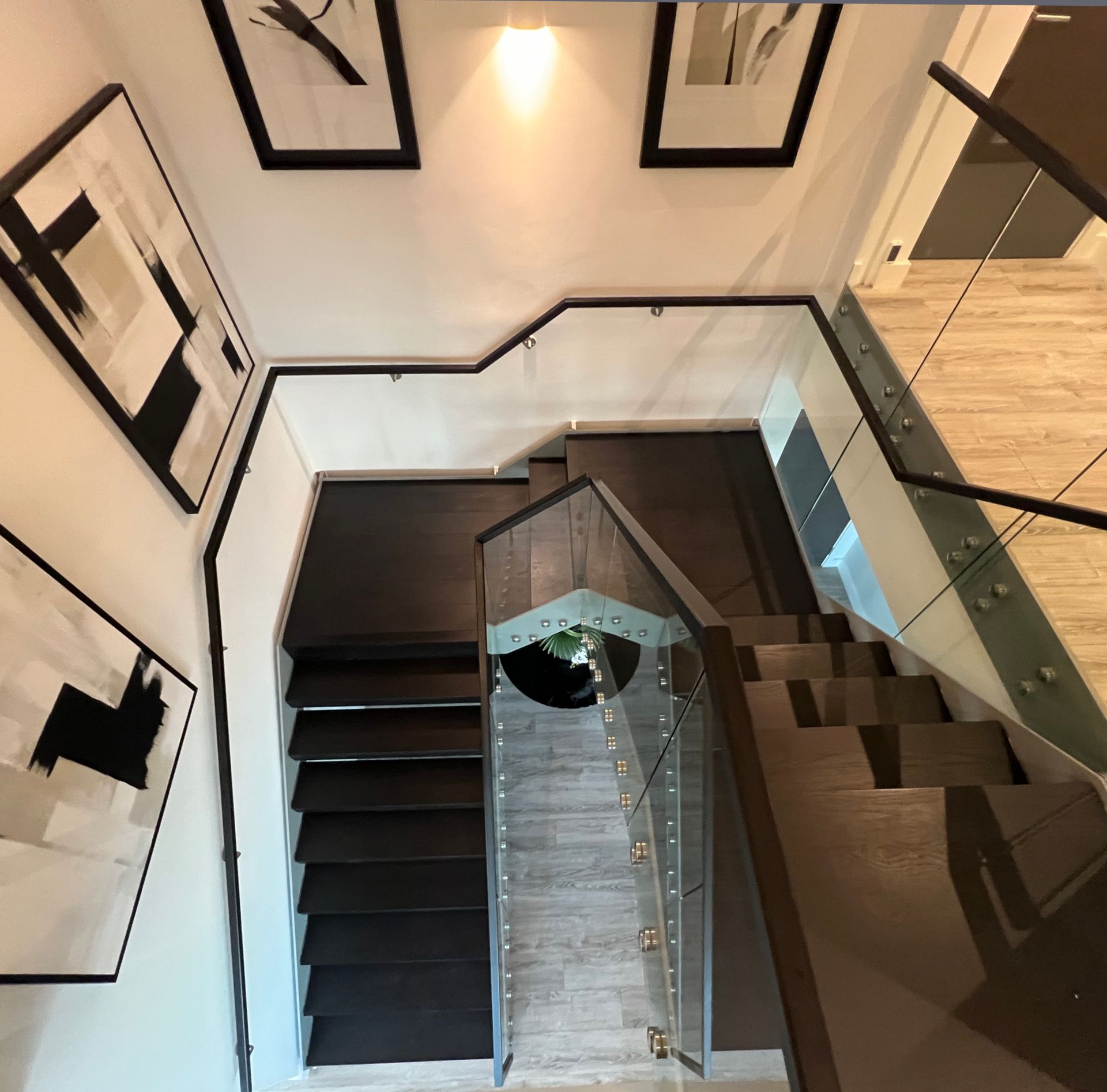 Looking down a modern staircase with dark wood steps, glass railing, and art on white walls.