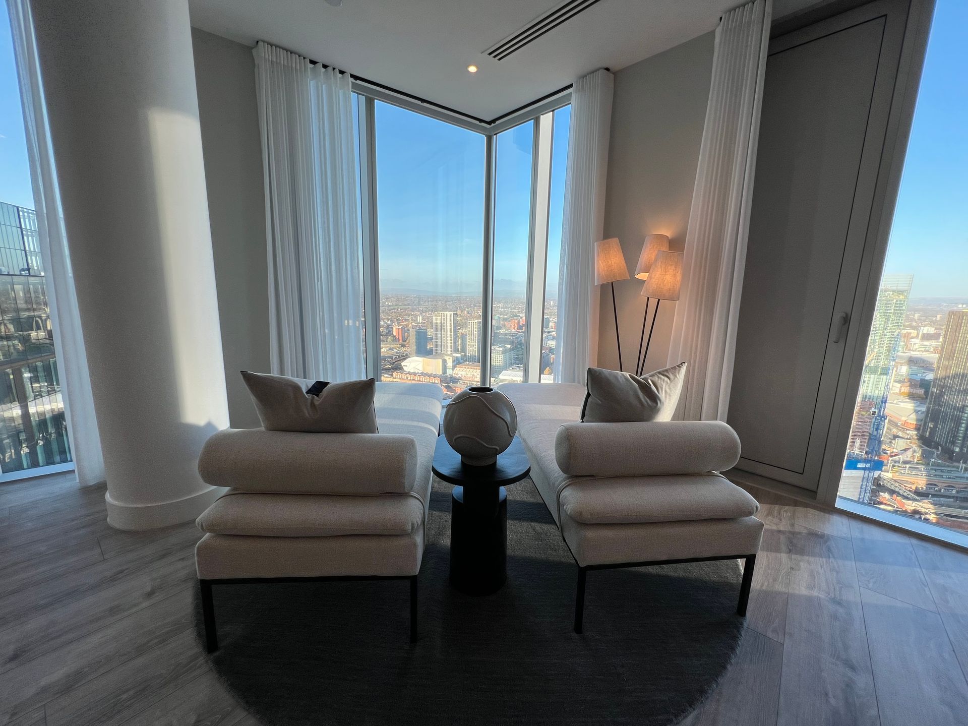 Two cream-colored armchairs face each other near a window with city views. A lamp and a decorative vase sit nearby.