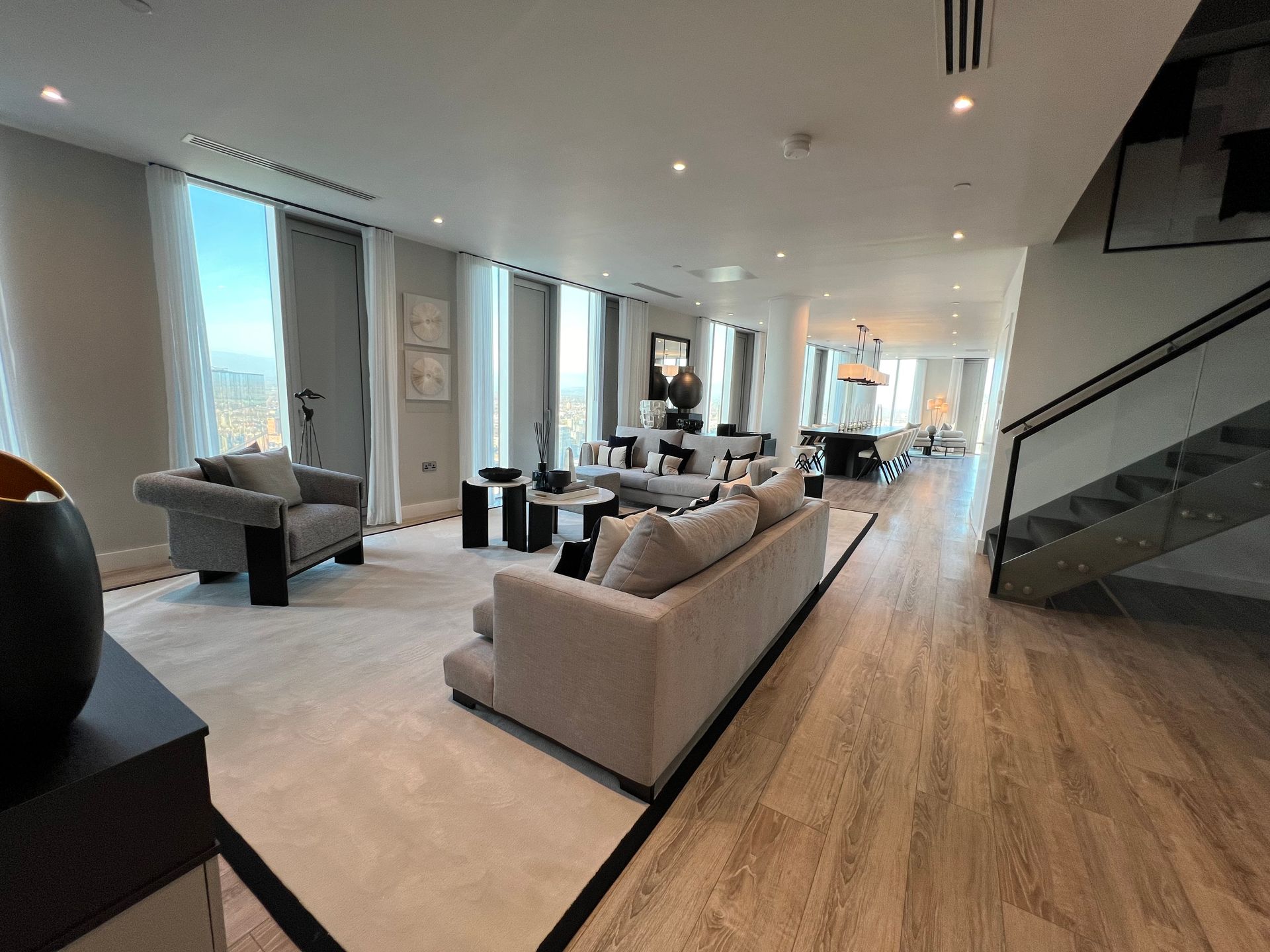 Spacious modern living room with floor-to-ceiling windows, neutral tones, and stairs leading upwards.
