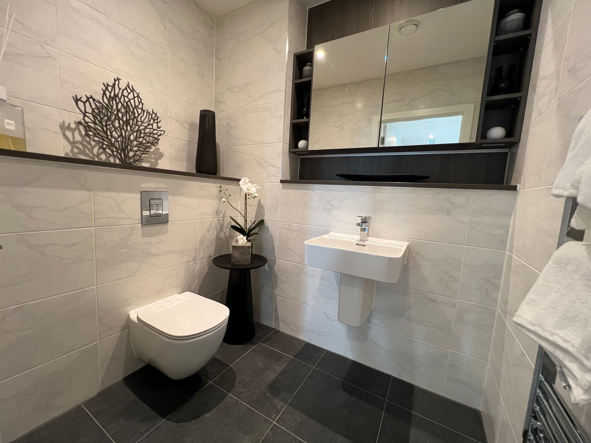 Modern bathroom with white walls, dark floor, and a floating toilet, sink, and shelf with decor.