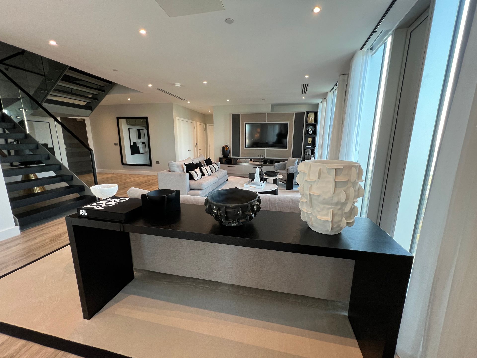 Living room with modern decor, black console table, gray sofa, stairs, and large windows.