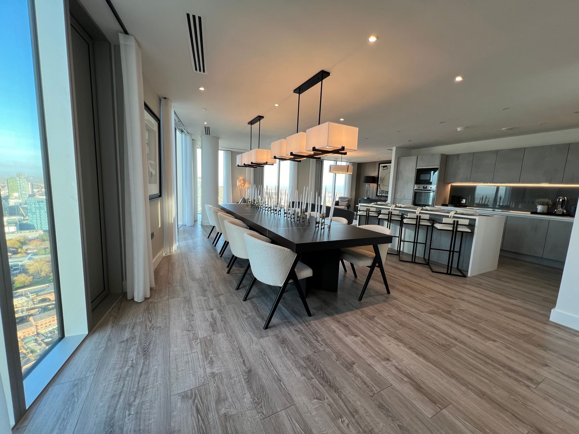 Spacious dining area with long table, chairs, and overhead lights. Kitchen with island and stools in the background.