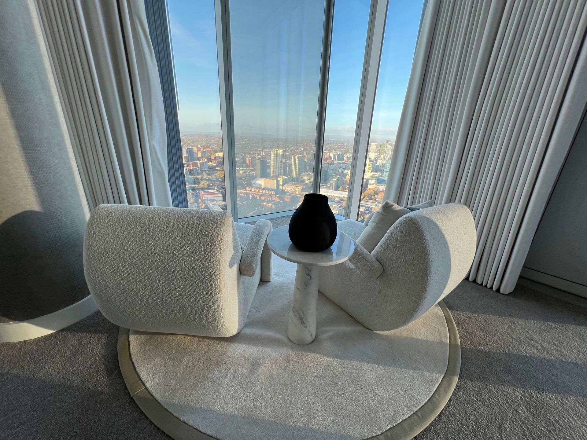 Two cream armchairs face a cityscape view through a large window. A small table holds a black vase on a round rug.