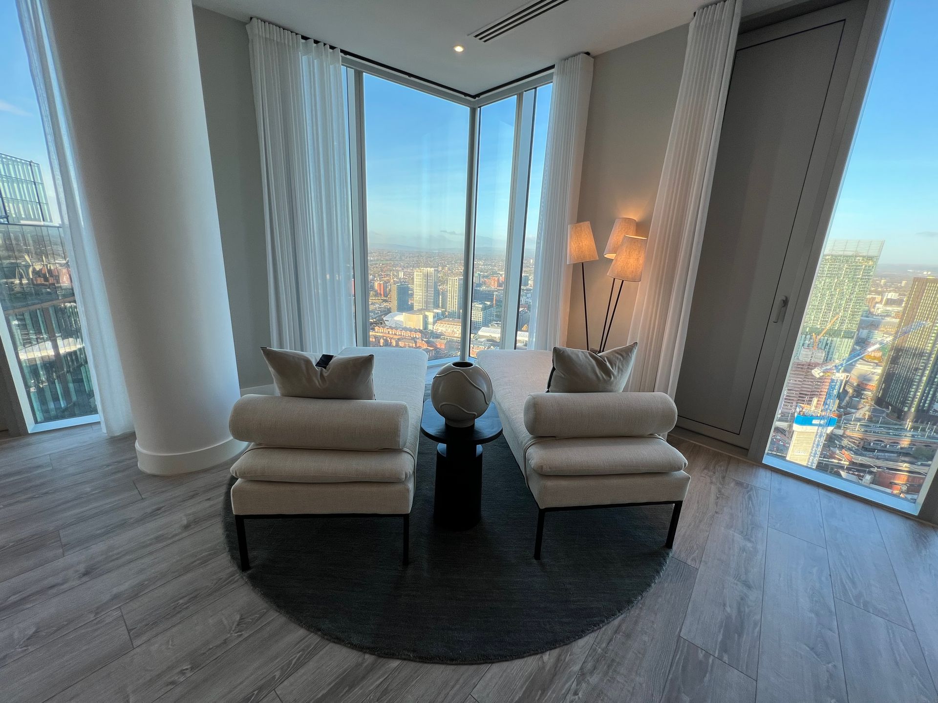 Two cream chairs face a cityscape, with a round rug and tall windows in a high-rise building.