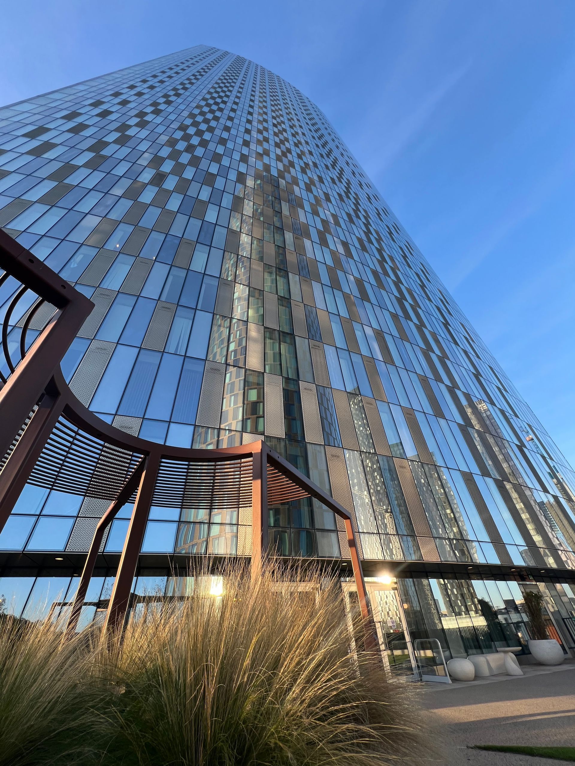Tall, modern glass skyscraper against a blue sky, foreground with a brown metal structure and dry grasses.