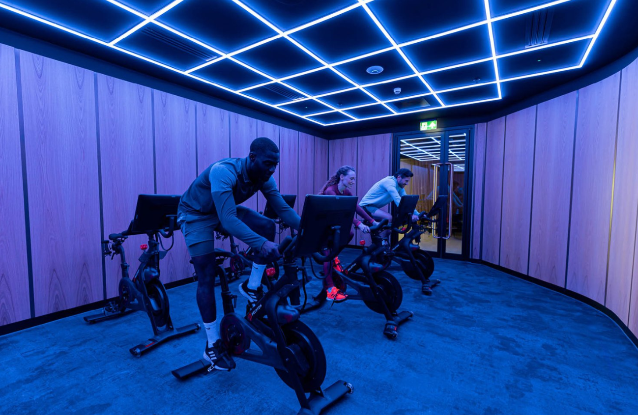 People cycling on stationary bikes in a room with blue lighting and wood-paneled walls.