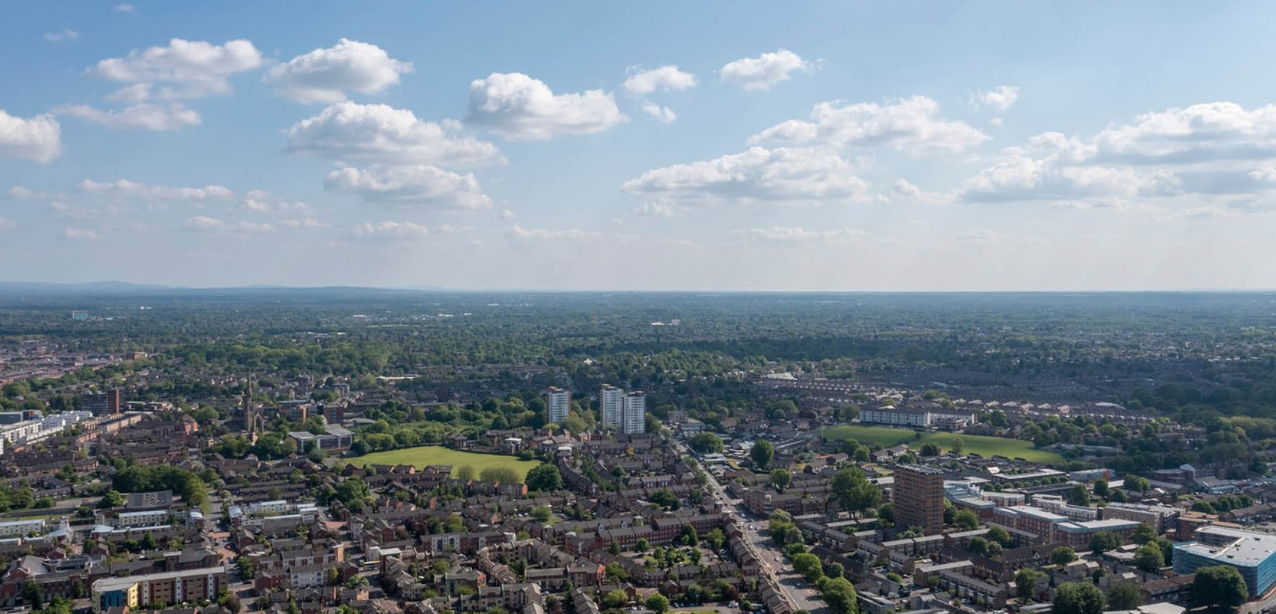 Aerial view of a city with buildings and green spaces under a blue sky with clouds.