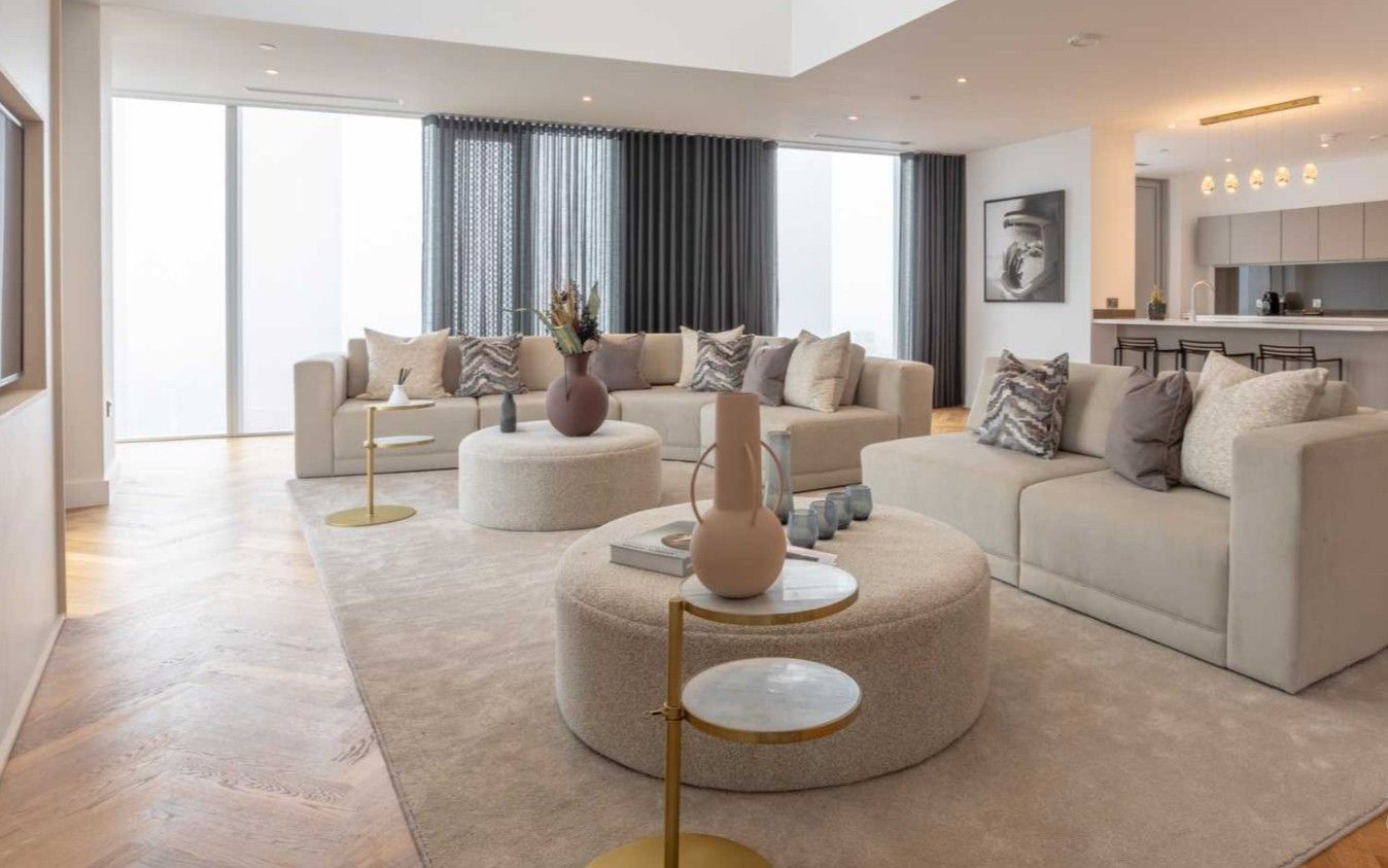 Spacious living room with neutral tones. Beige sectional sofa, round ottoman, large windows, and hardwood floors.