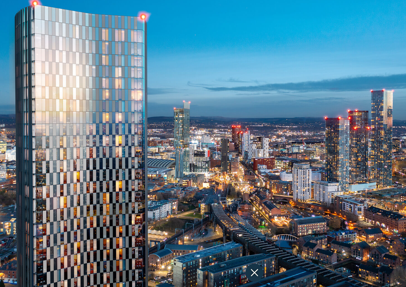 High-rise building dominates cityscape at dusk with city lights. Other tall buildings and railway tracks also visible.
