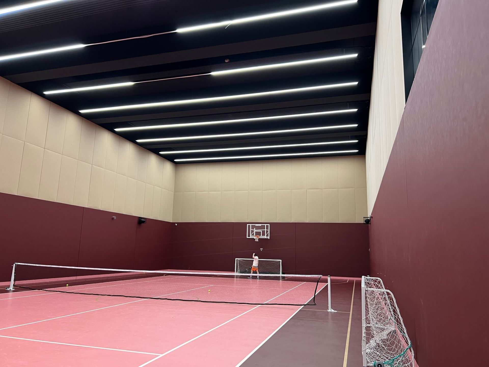 Indoor sports court with red flooring and walls, and white ceiling lights. A net and basketball hoop are visible.