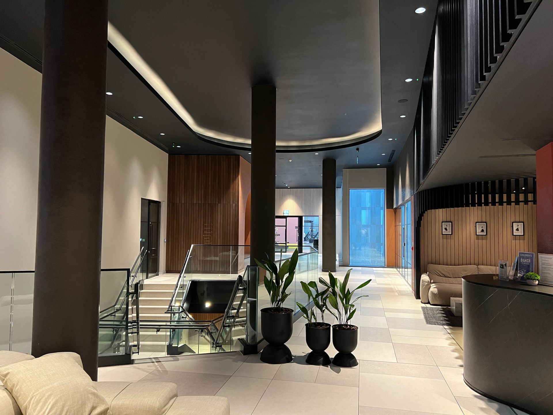 Lobby with staircase, plants, seating, and a front desk. Modern design with dark ceiling and wood paneling.