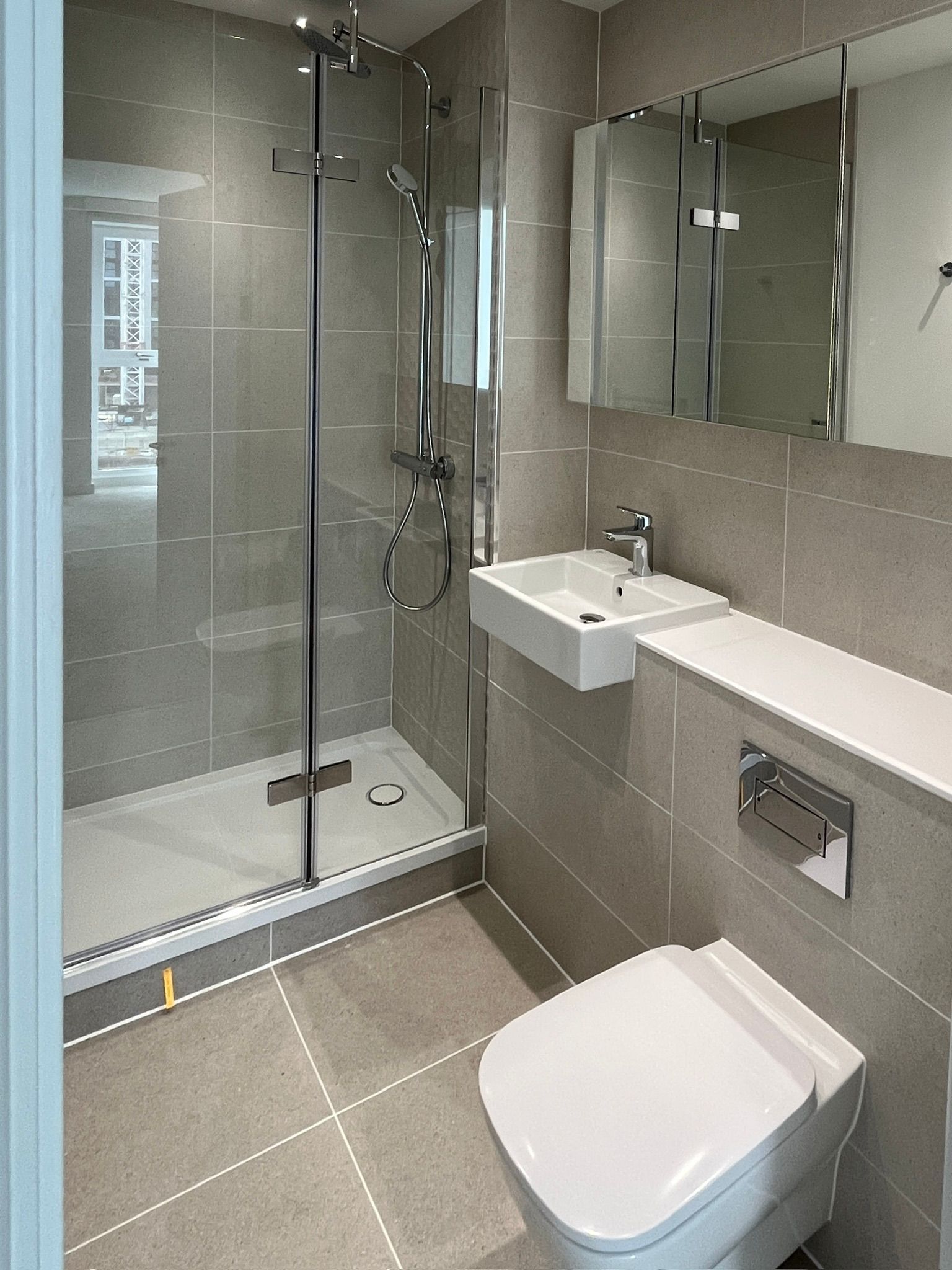 Modern bathroom with a shower, sink, toilet, and mirror. Gray tiles, chrome fixtures, white fixtures.