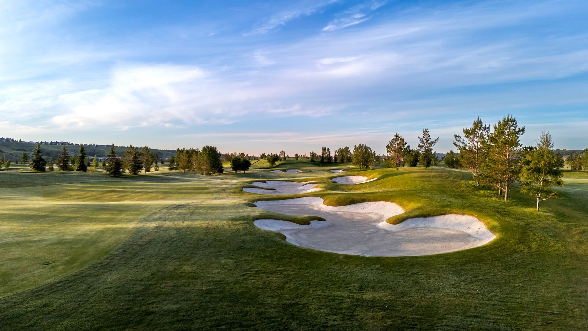 Golf course fairway with sand traps, green grass, trees, and a blue sky.