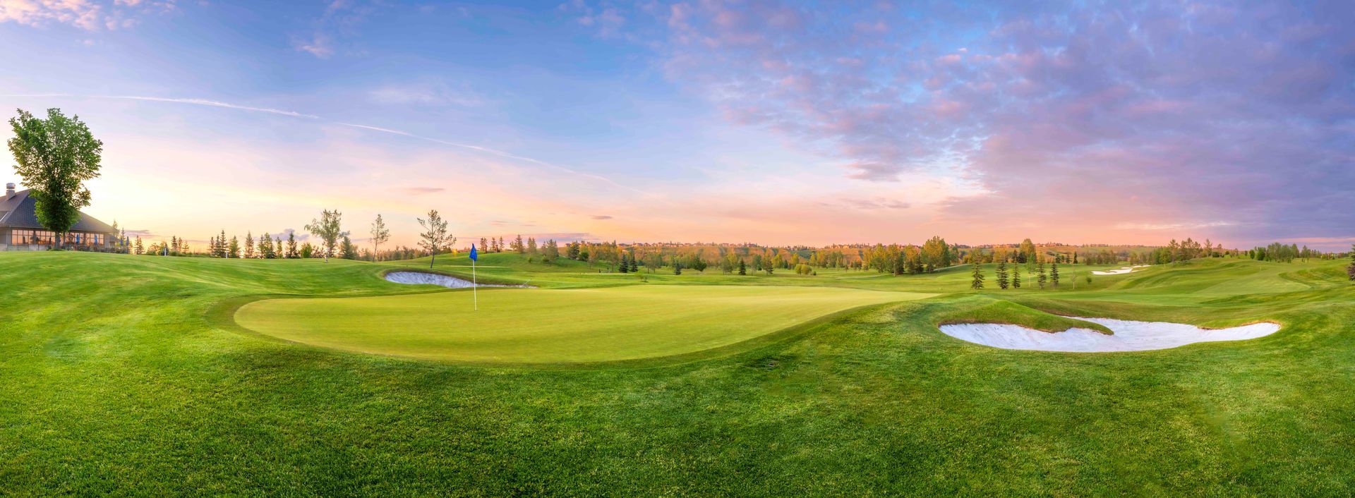 Golf green with blue flag, trees, and blue sky.