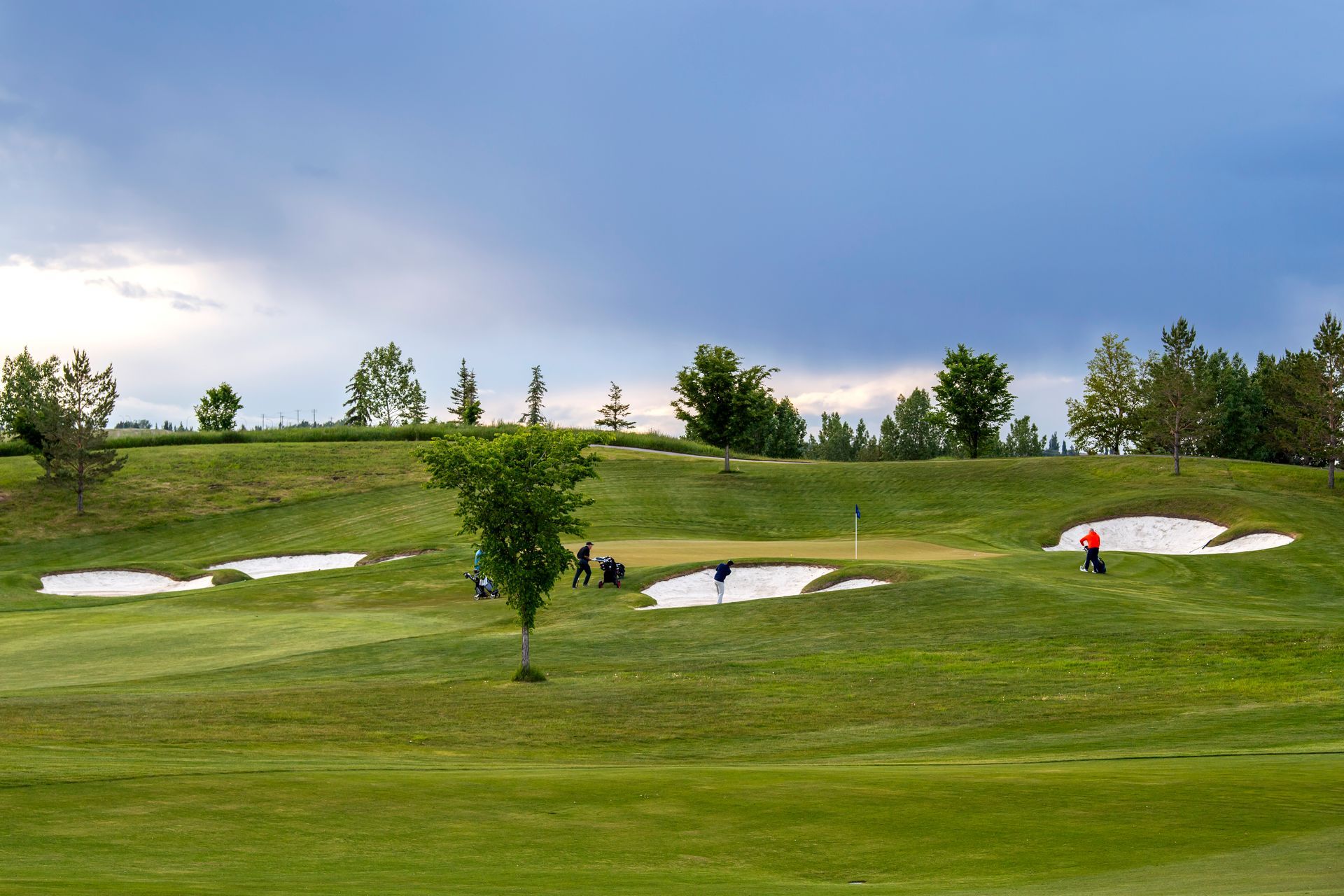 Golf course scene: players on green near sand traps, under a cloudy sky.