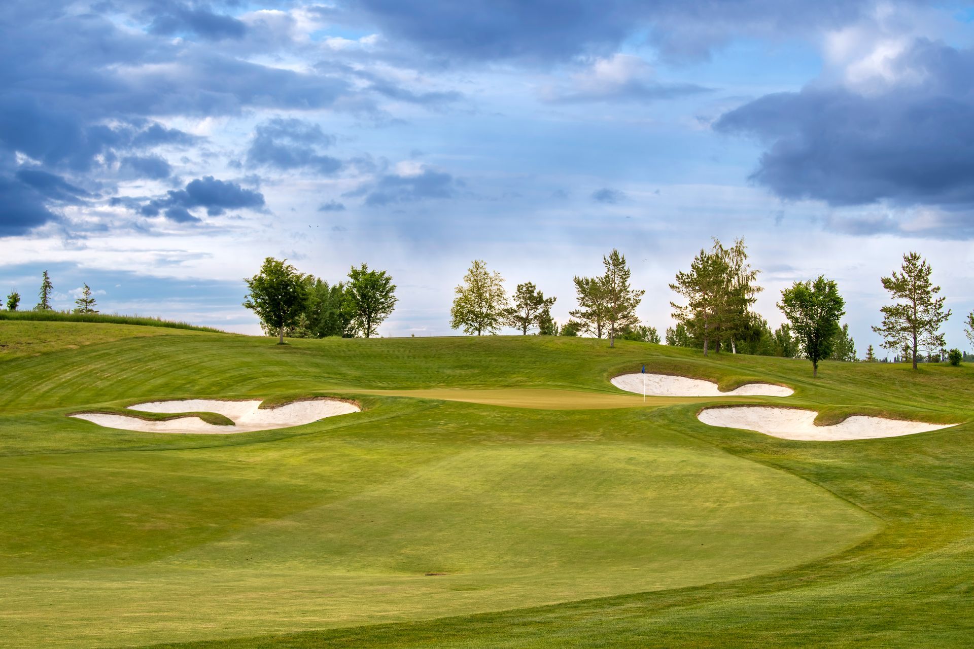 Golf course green with sand traps, trees, and cloudy sky.