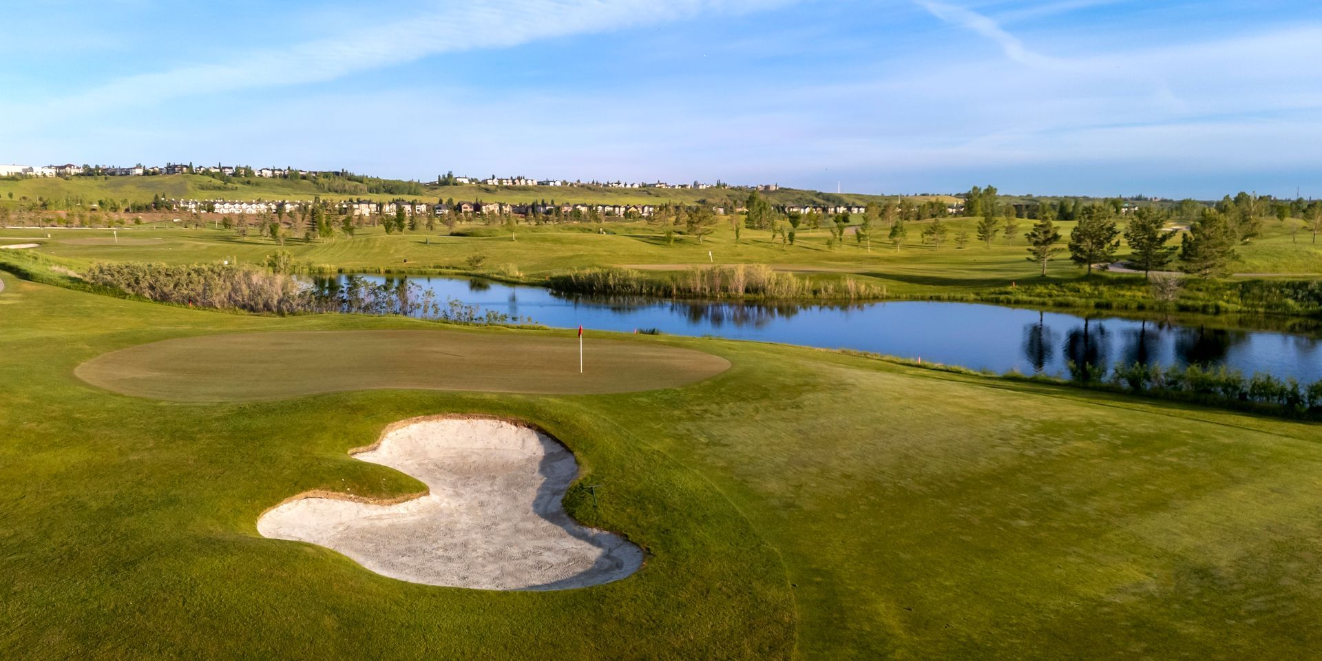 A golf course with a sand trap, green, and water feature under a blue sky.
