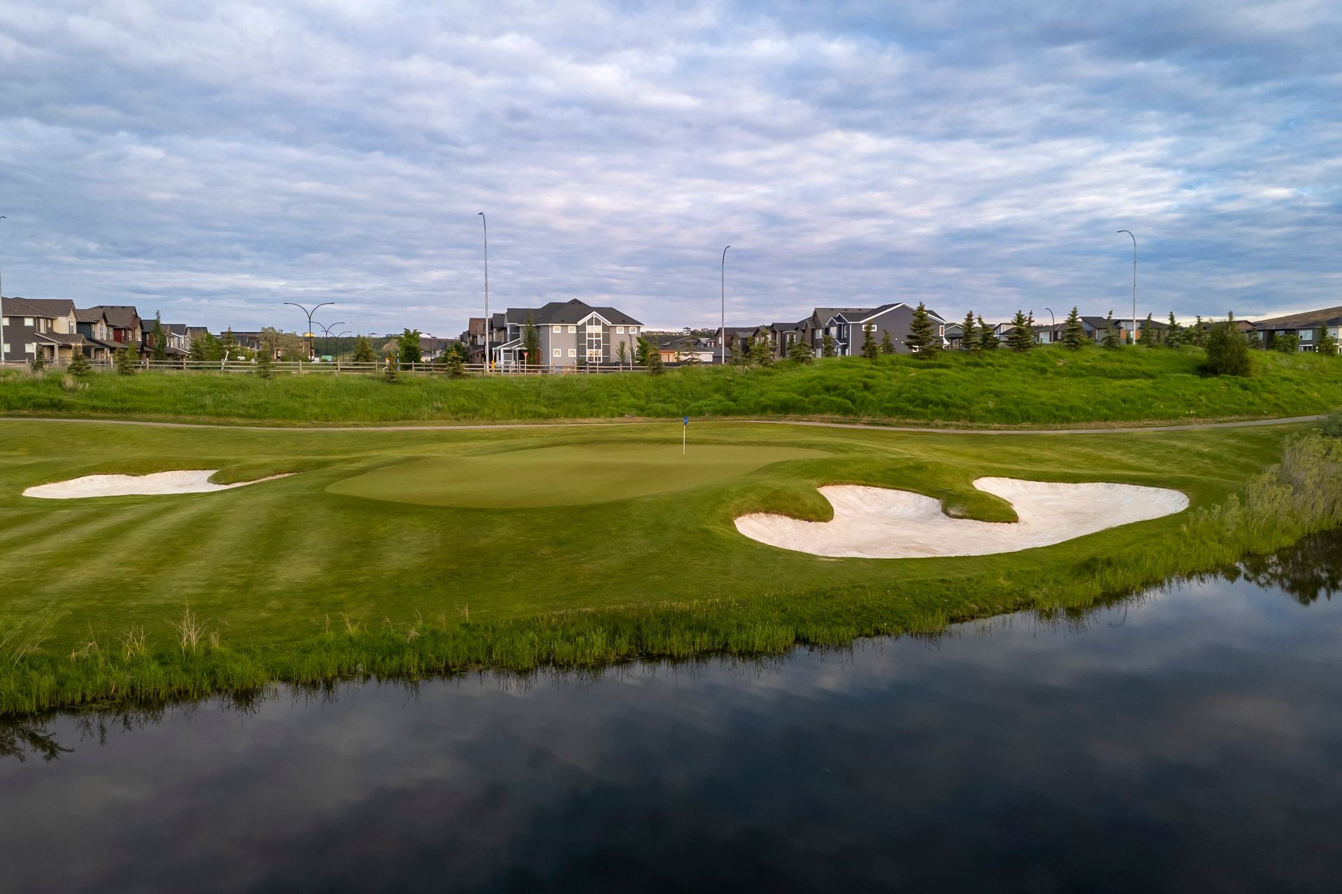 Golf course green with bunkers, water hazard, and houses in background under cloudy sky.