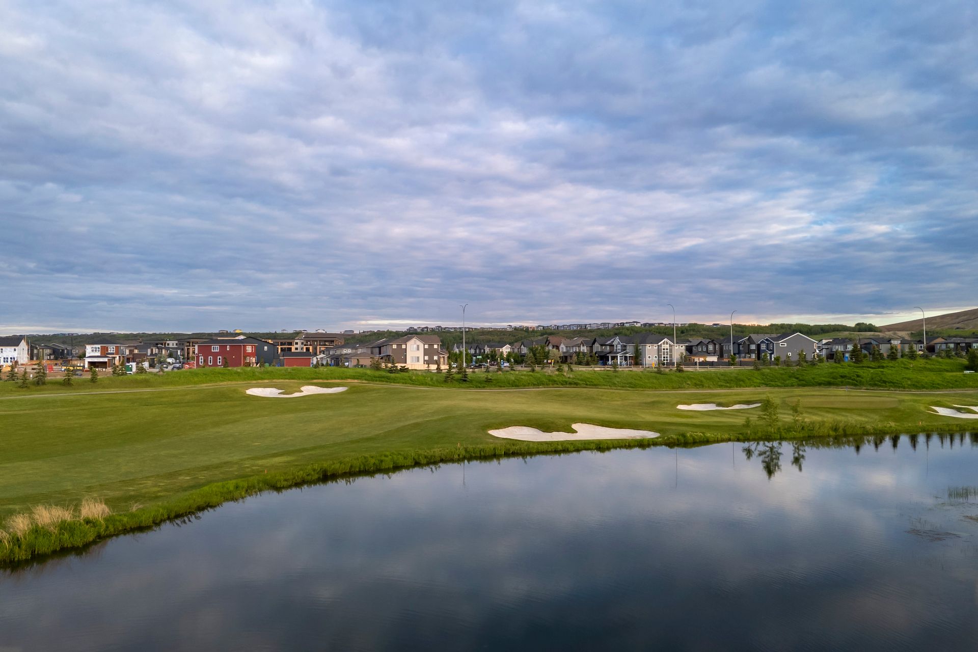 Golf course with houses in the background under a cloudy sky, water in the foreground.