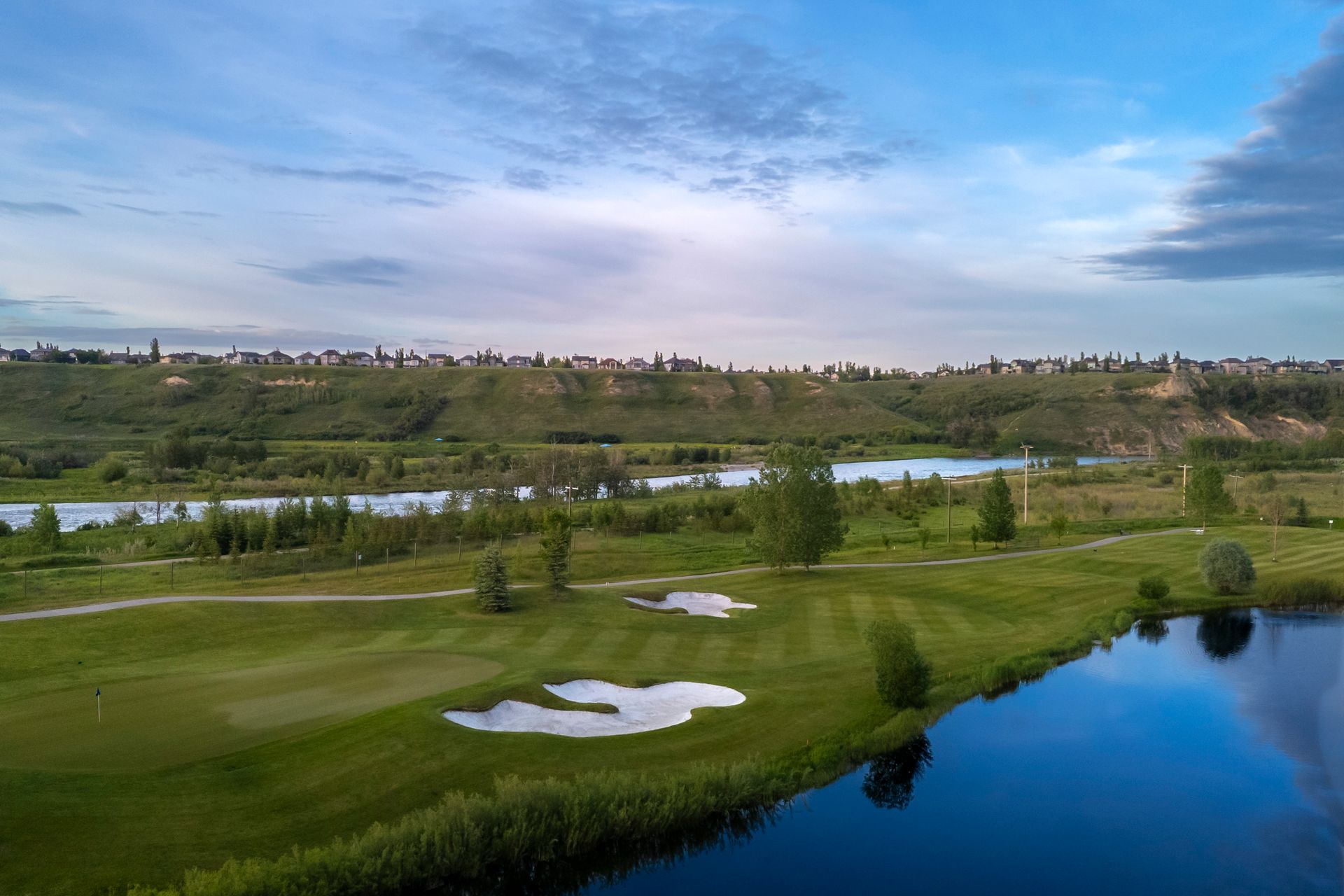 Aerial view of a golf course with green grass, sand traps, and a blue lake. Hills and a cloudy sky in the background.