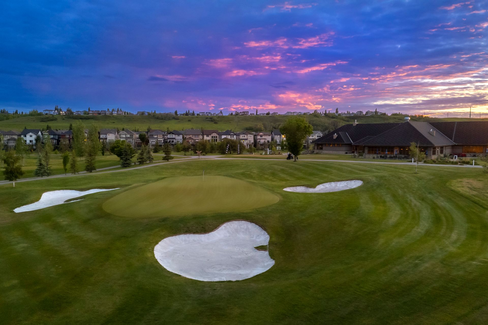 Green golf course with sand traps and clubhouse under a vibrant sunset sky.