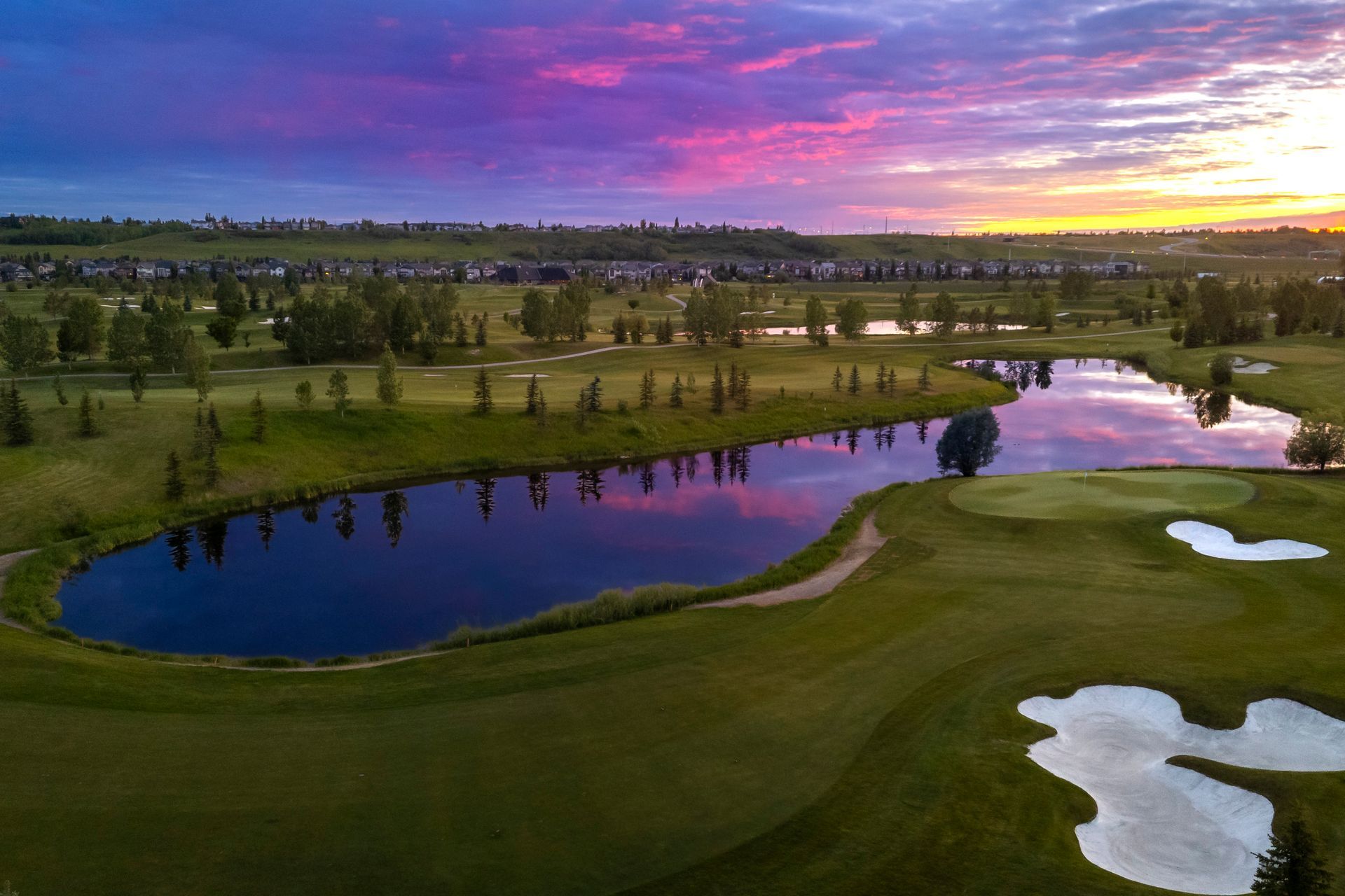Golf course with pond reflecting vibrant sunset sky. Green fairways, trees, and sand traps.