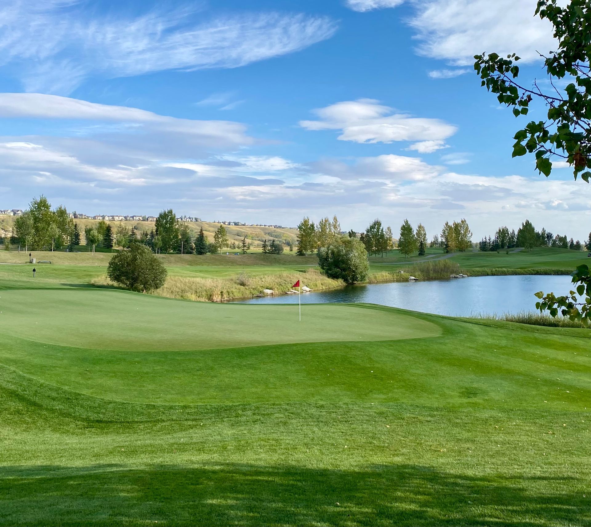 Golf course green with water feature, red flag, and blue sky with clouds.