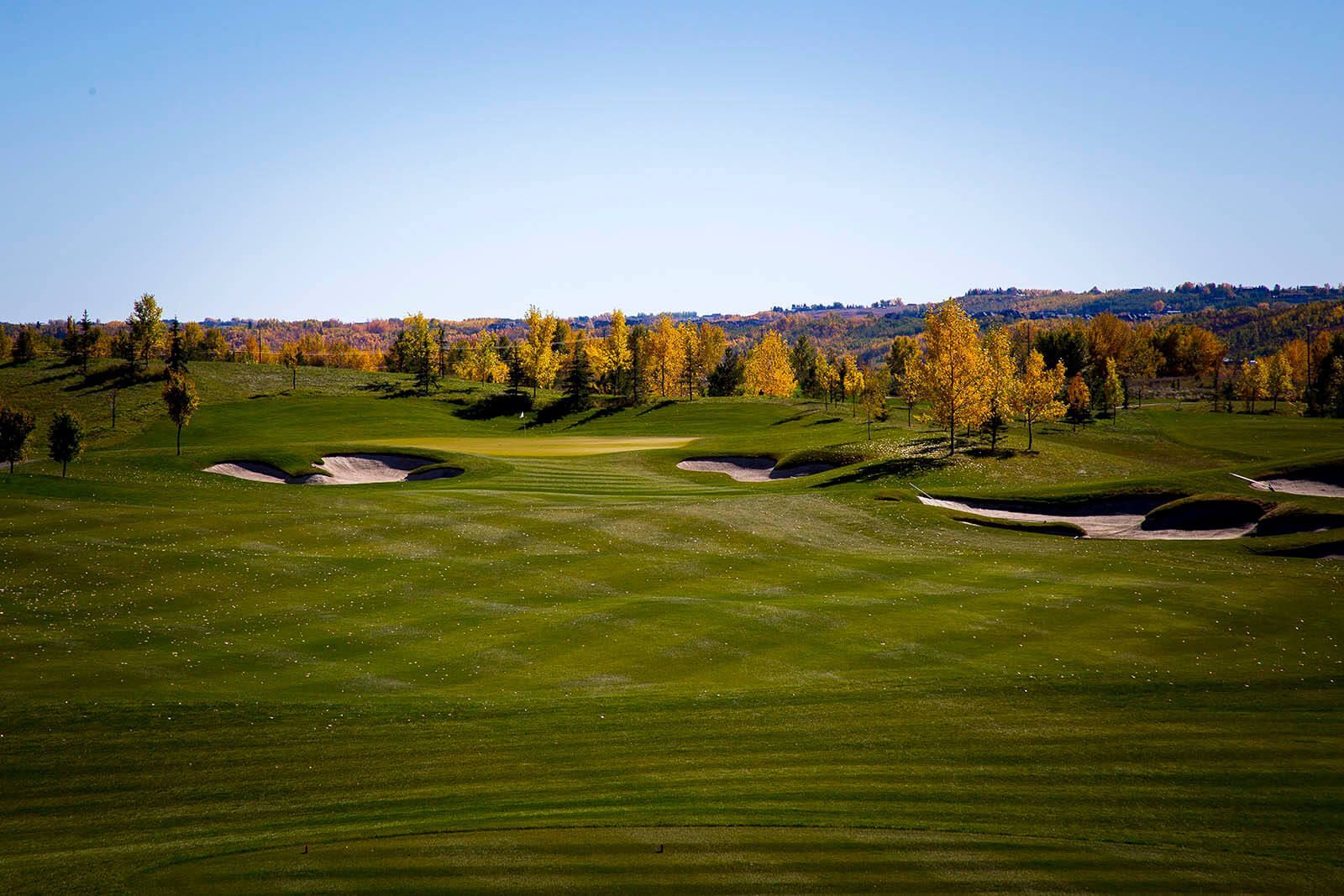 Golf course with green grass, sand traps, and trees with yellow fall foliage under a blue sky.