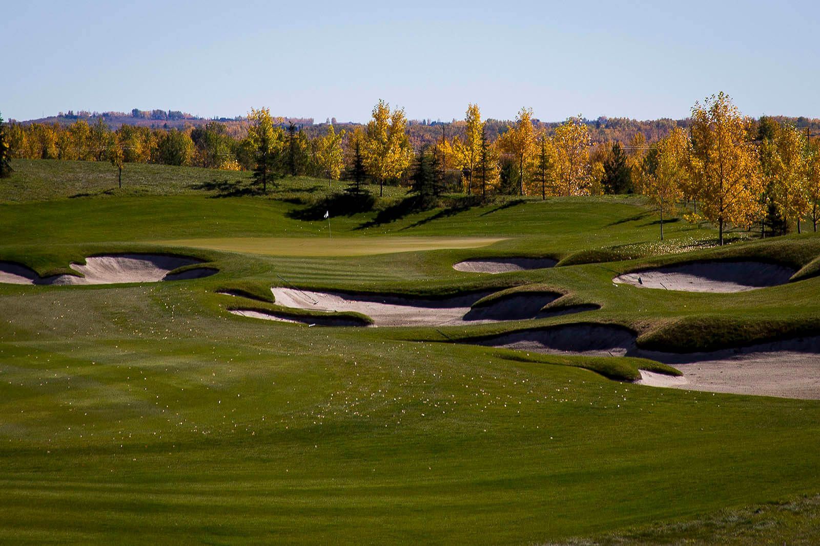 Golf course with green grass, sand bunkers, and trees with yellow leaves under a blue sky.