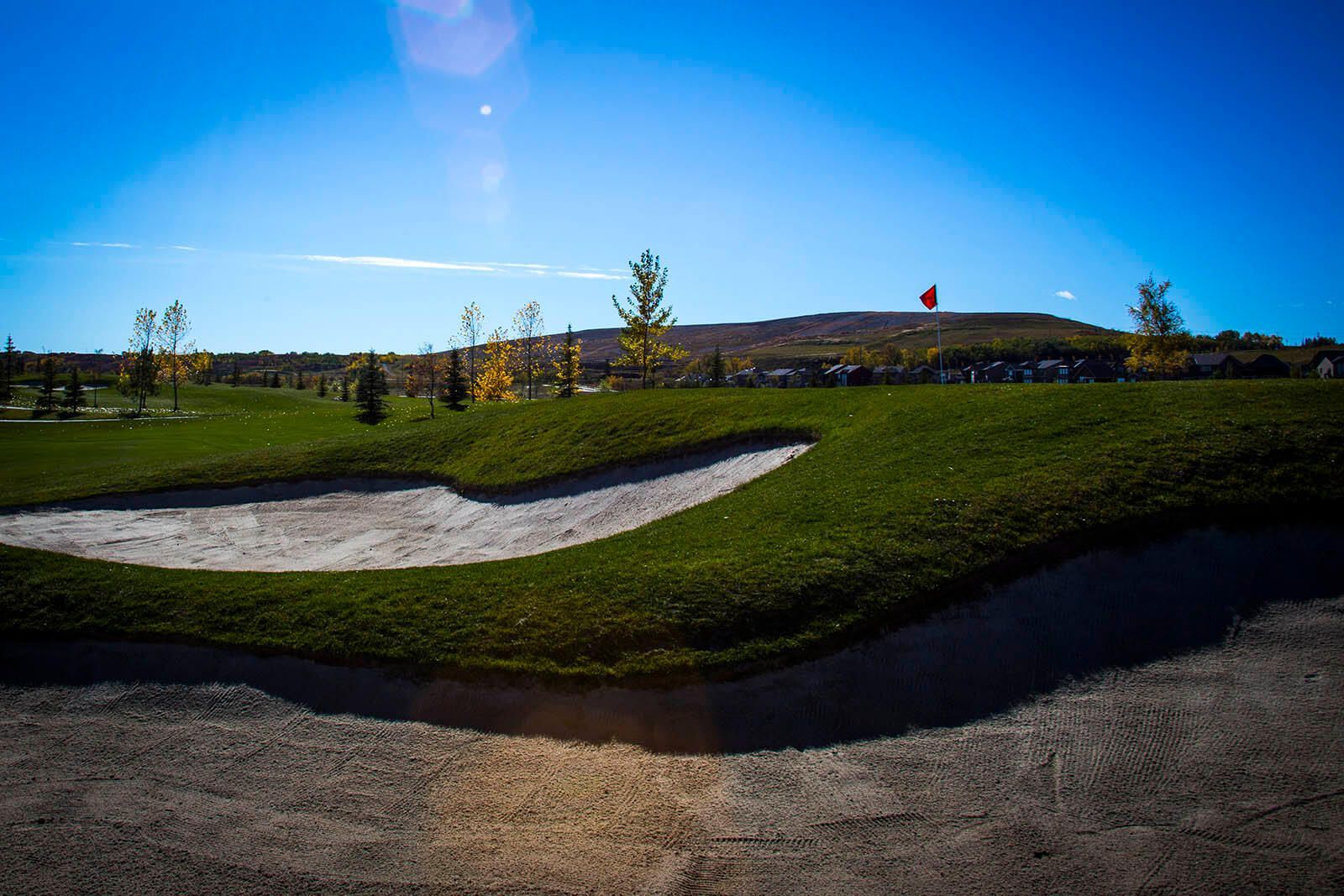 Golf course scene with green grass, sand trap, and distant buildings under a bright blue sky.
