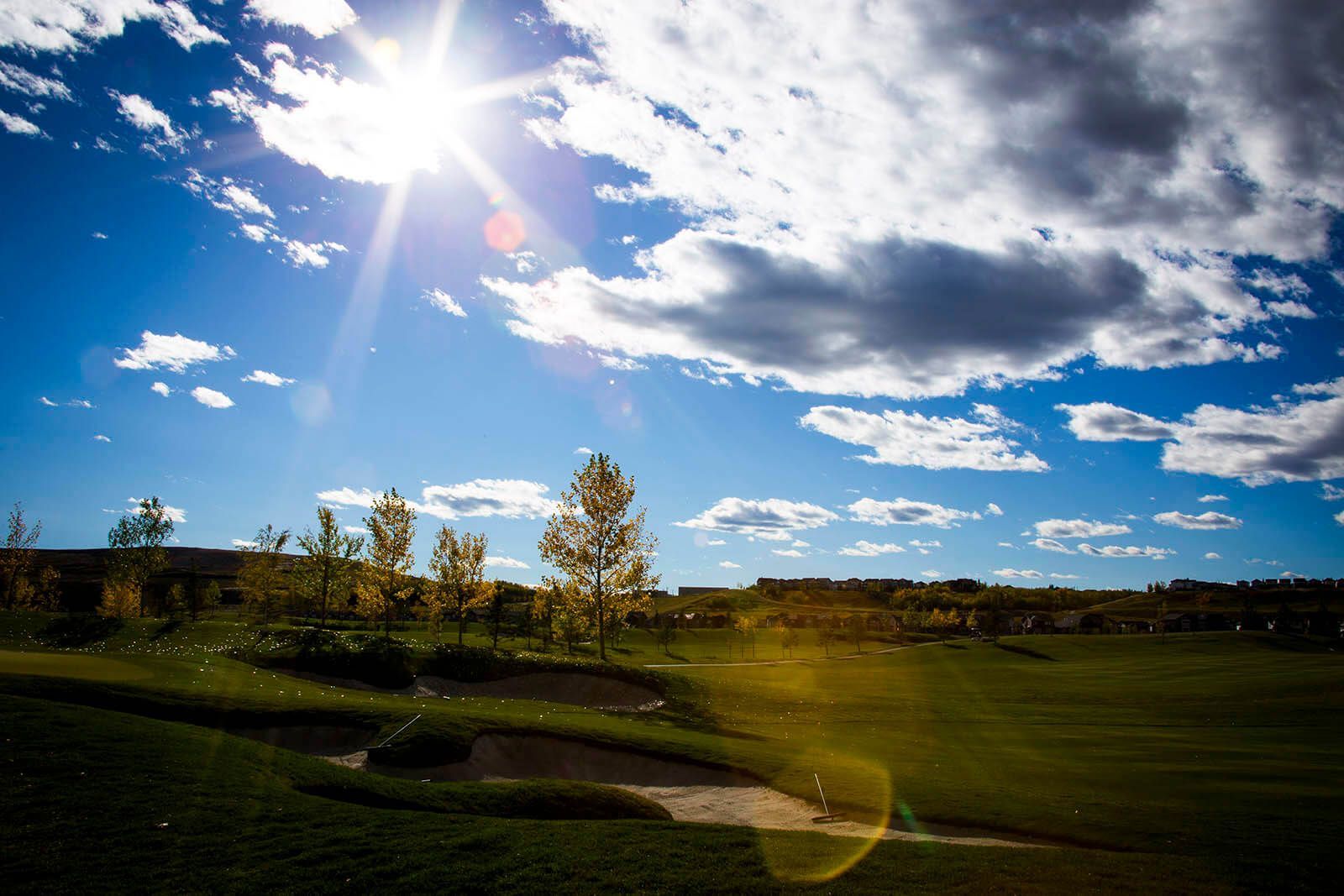 Bright sun over a golf course with green grass, trees, and cloudy blue sky.