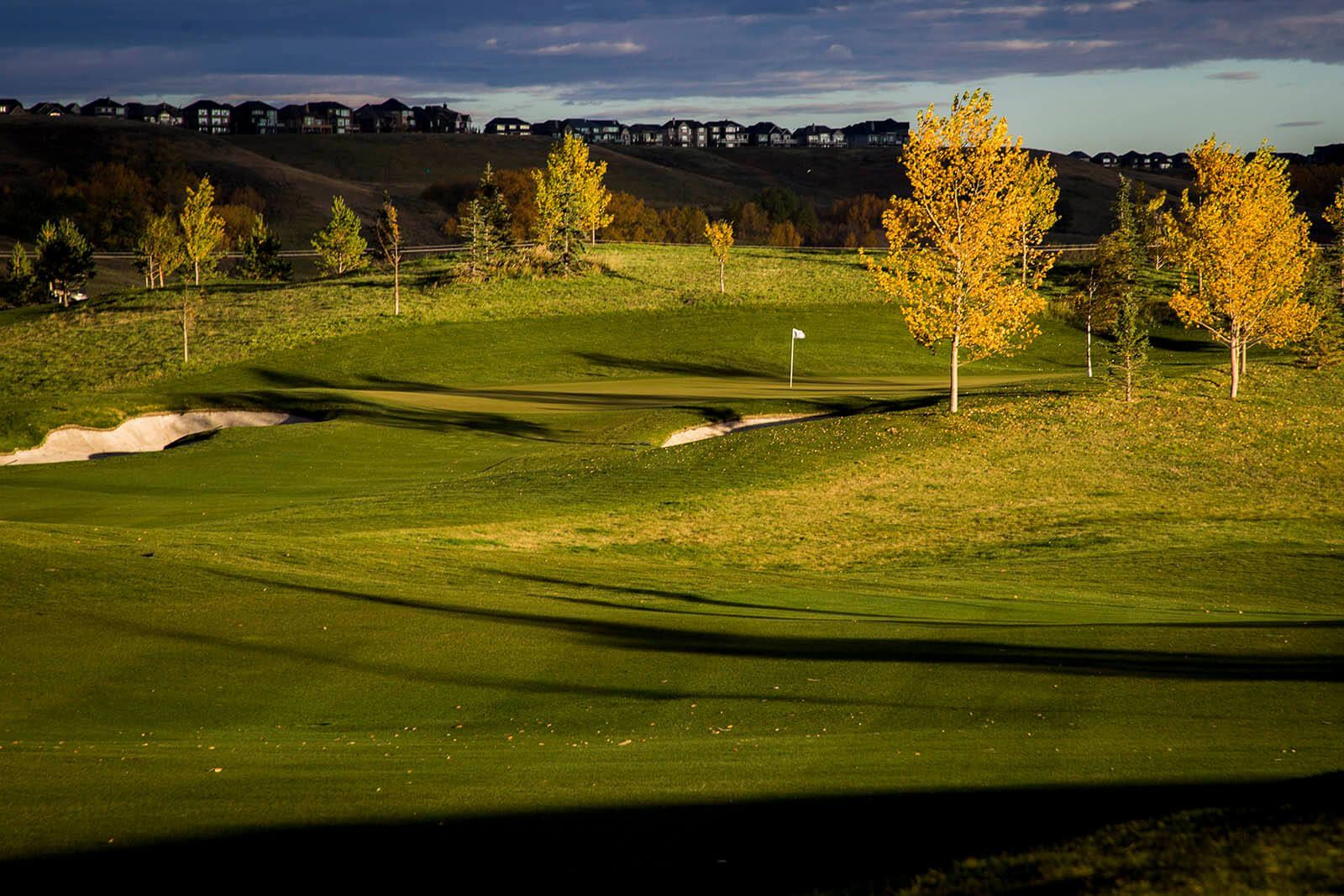 Golf course green with sand trap, trees in fall colors, houses on a distant hill.